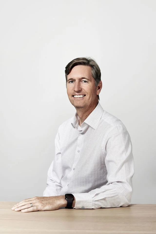 Portrait of a middle-aged man with short, dark hair, wearing a white button-up shirt and a smartwatch, sitting at a wooden table against a plain white background.