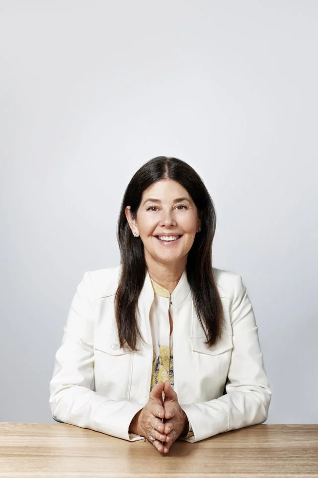 A woman with long dark hair wearing a white blazer sitting at a light wooden table, smiling, with a plain white background.