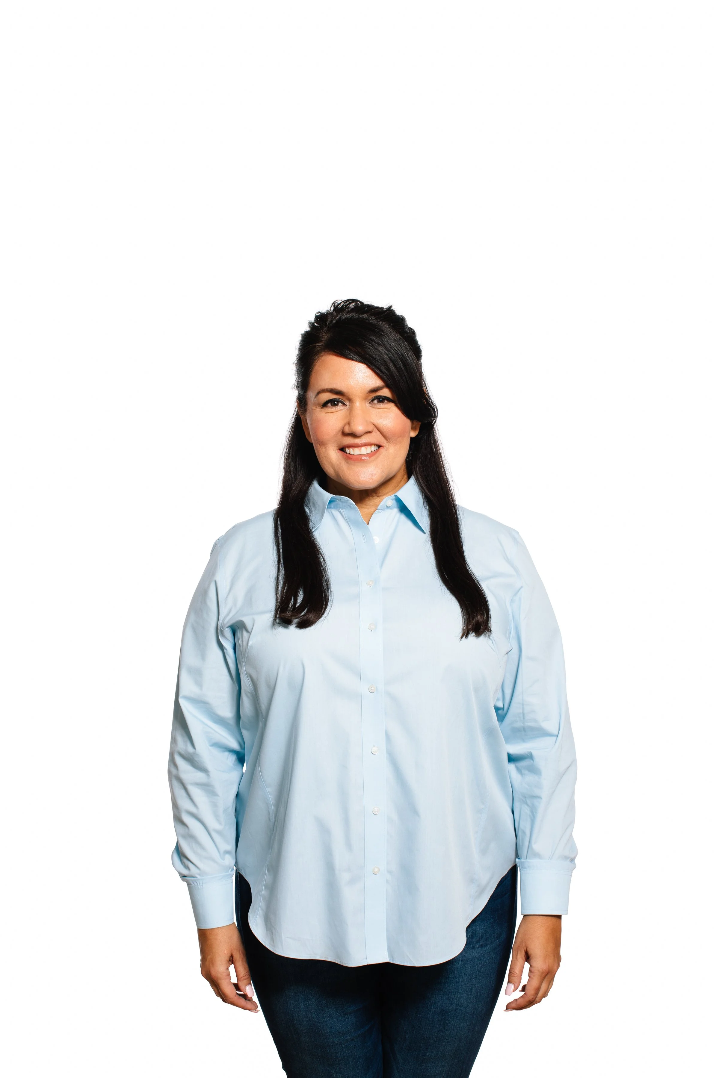 A woman with long black hair wearing a light blue button-up shirt and dark jeans, smiling at the camera against a plain white background.