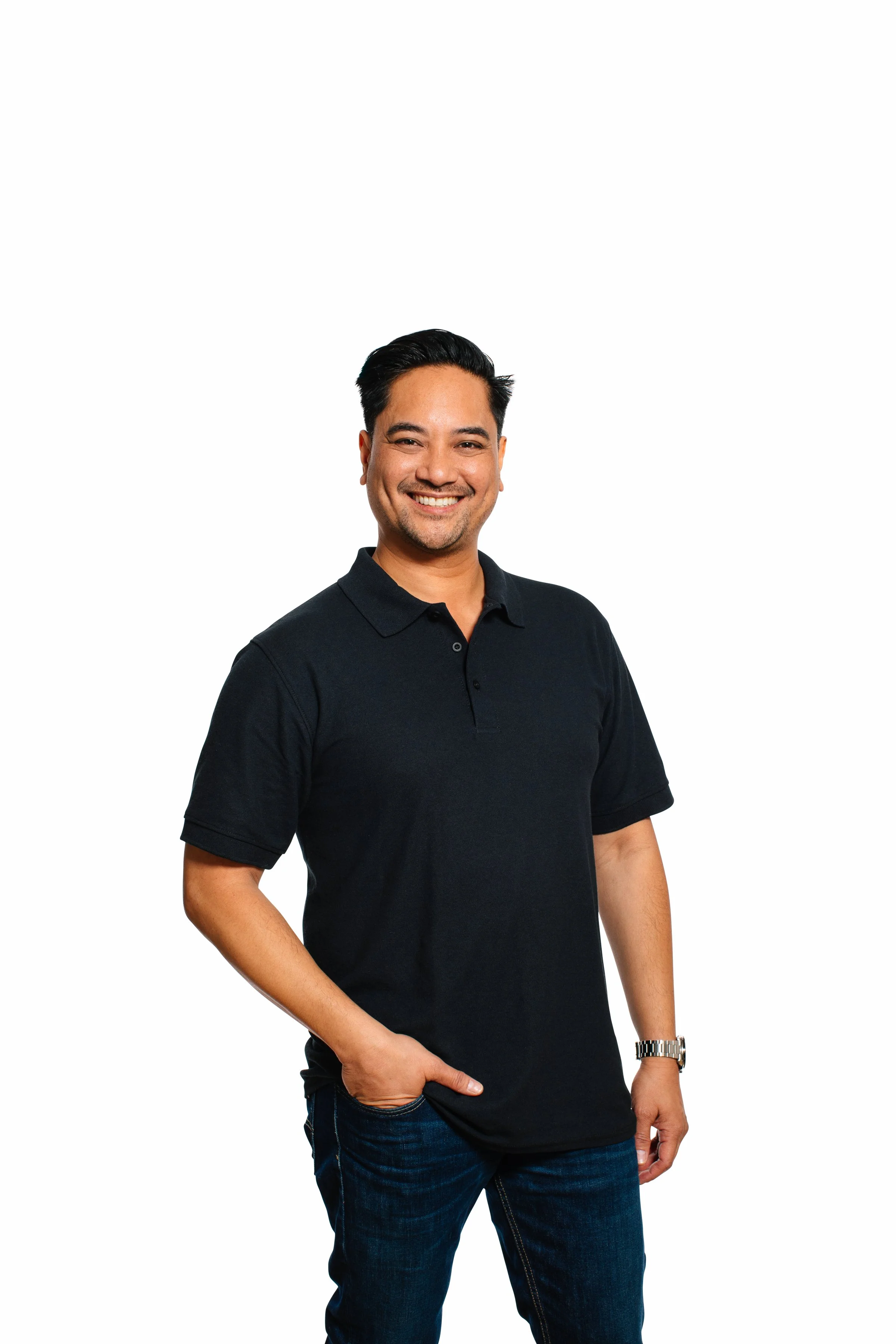 A smiling man with short dark hair, wearing a black polo shirt and dark jeans, standing against a plain white background.