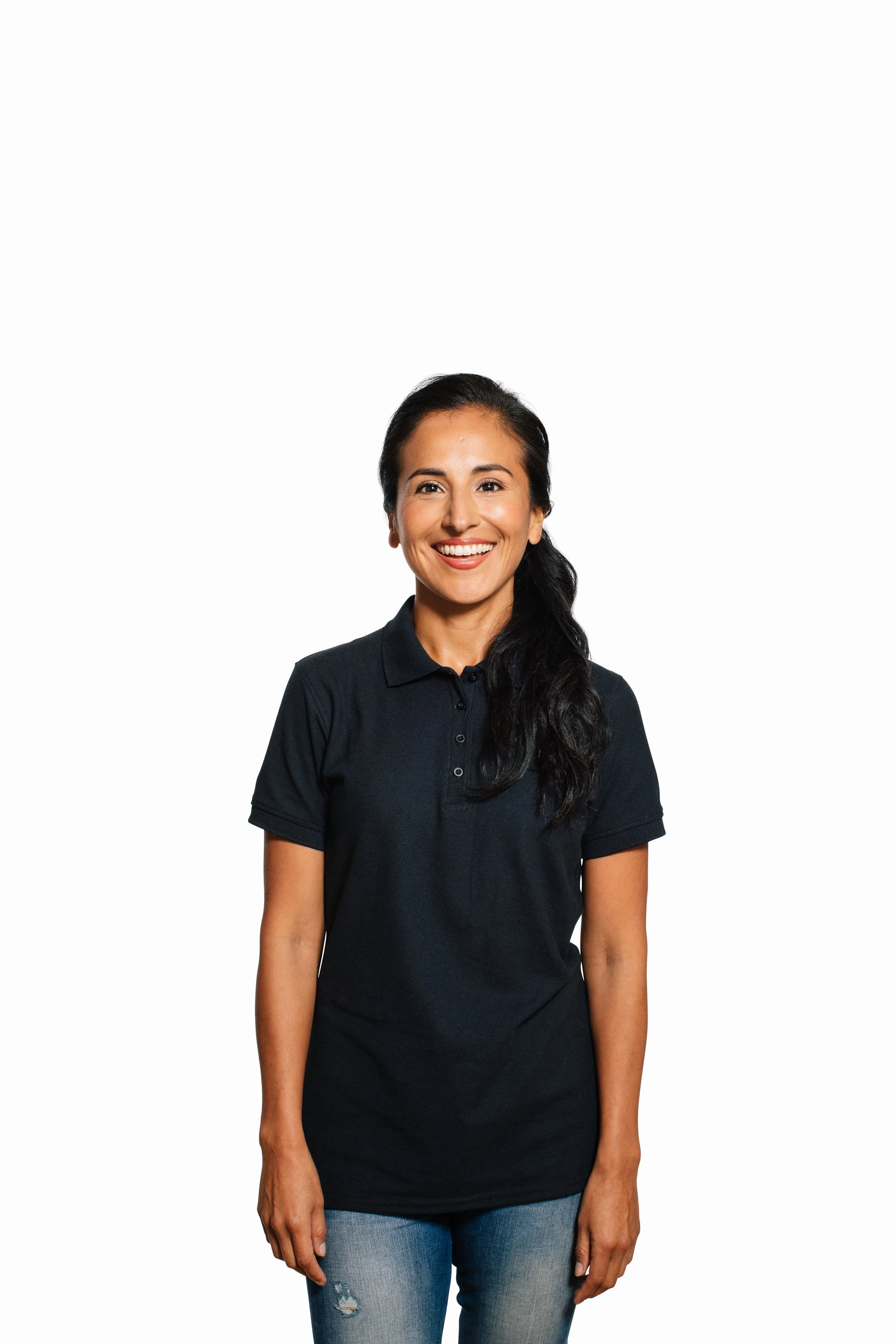 A smiling woman with long dark hair, wearing a black polo shirt and ripped jeans, standing against a plain white background.