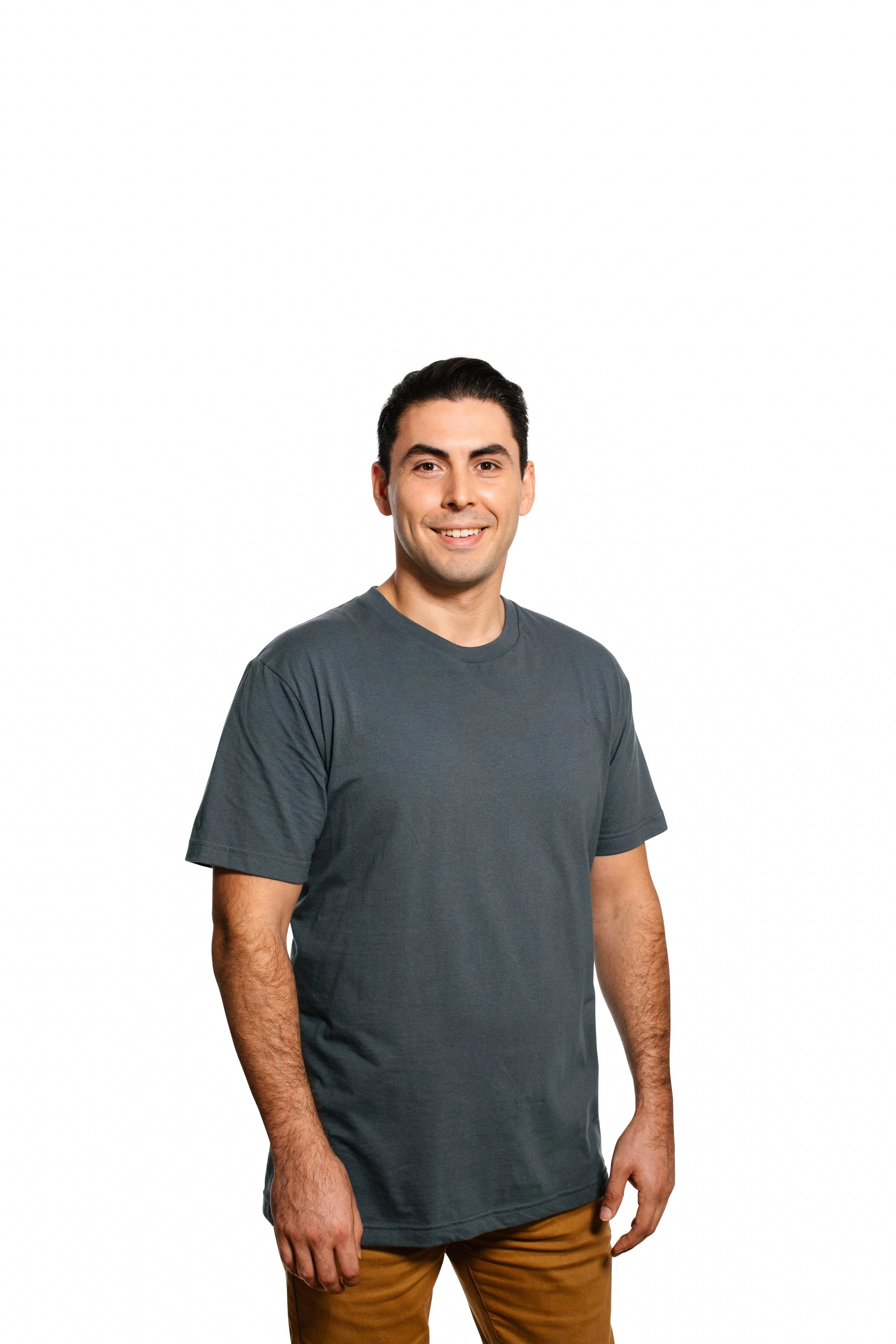 A man wearing a dark gray t-shirt and light brown pants standing against a plain white background.