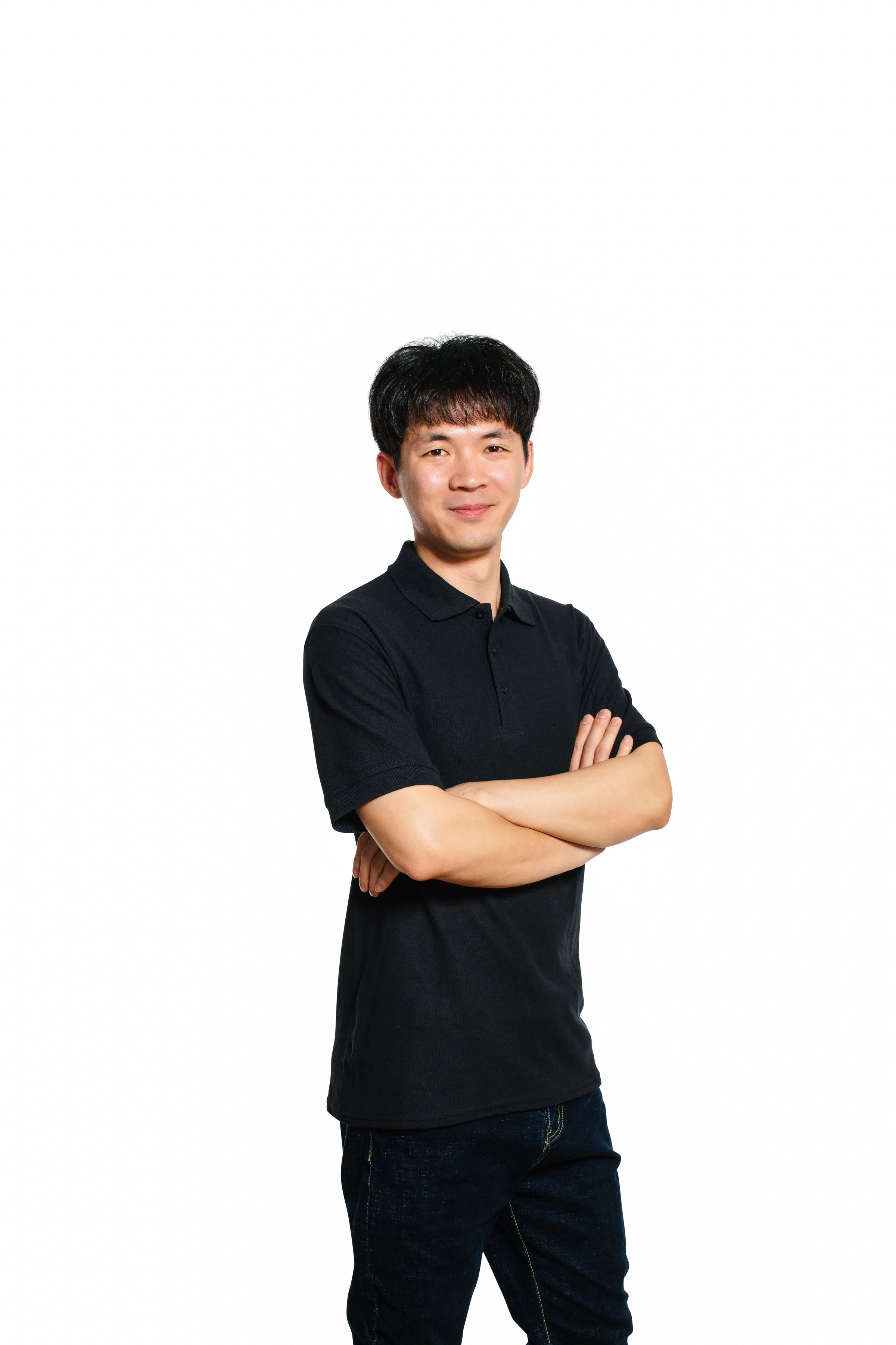 A young man in a black polo shirt with arms crossed, standing against a white background, smiling slightly.