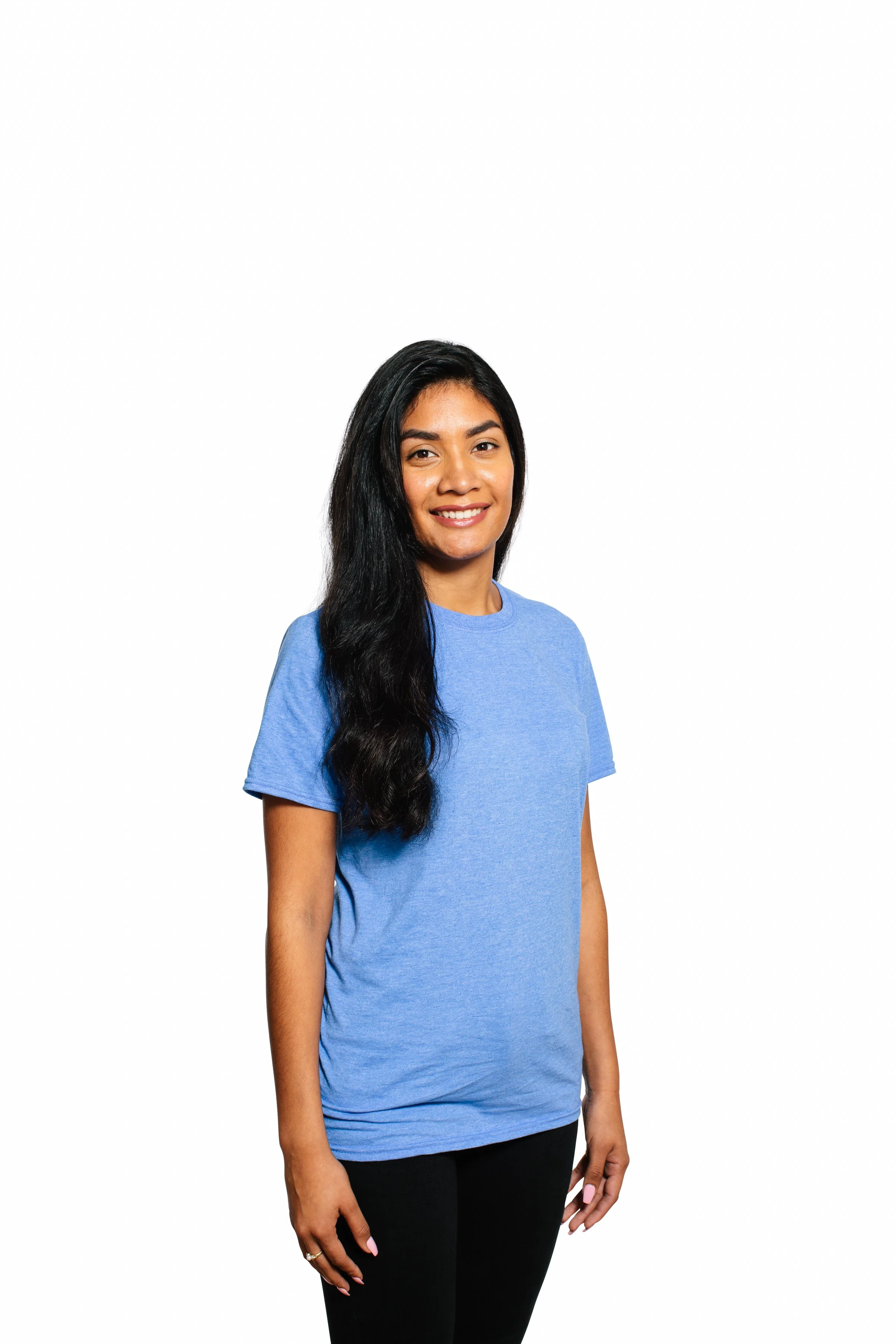A smiling woman with long black hair wearing a blue t-shirt and black pants standing against a plain white background.