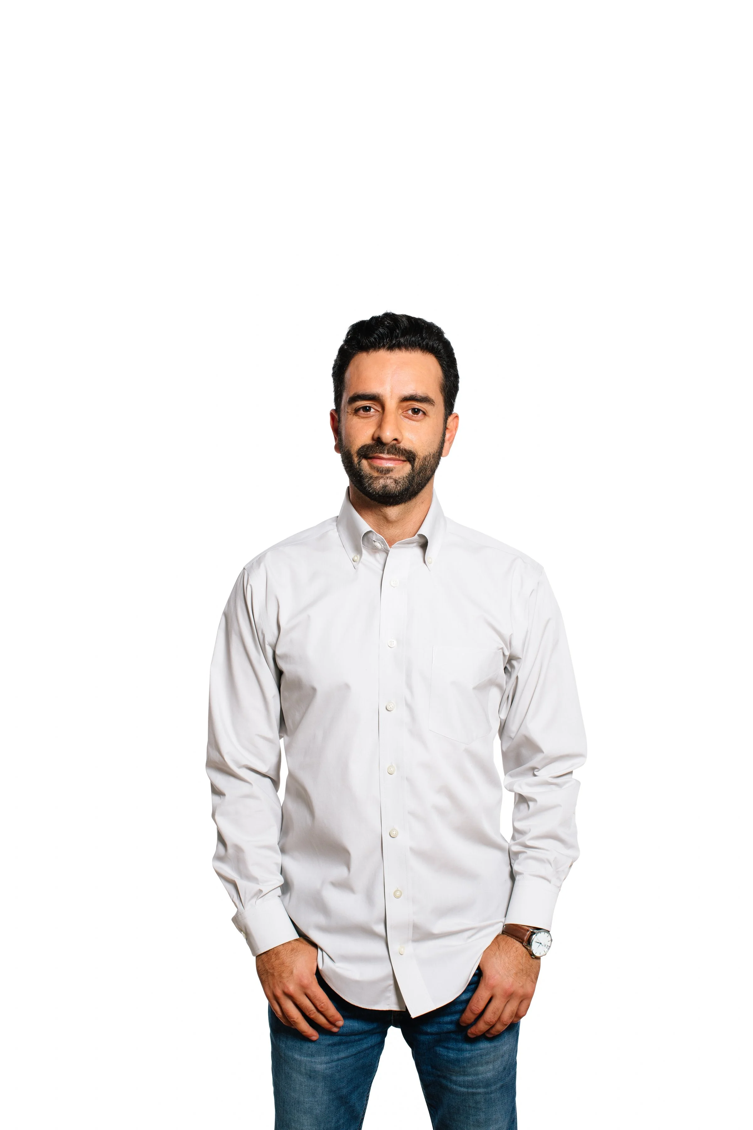 A man with dark hair, a beard, wearing a white button-up shirt and jeans, standing against a white background.