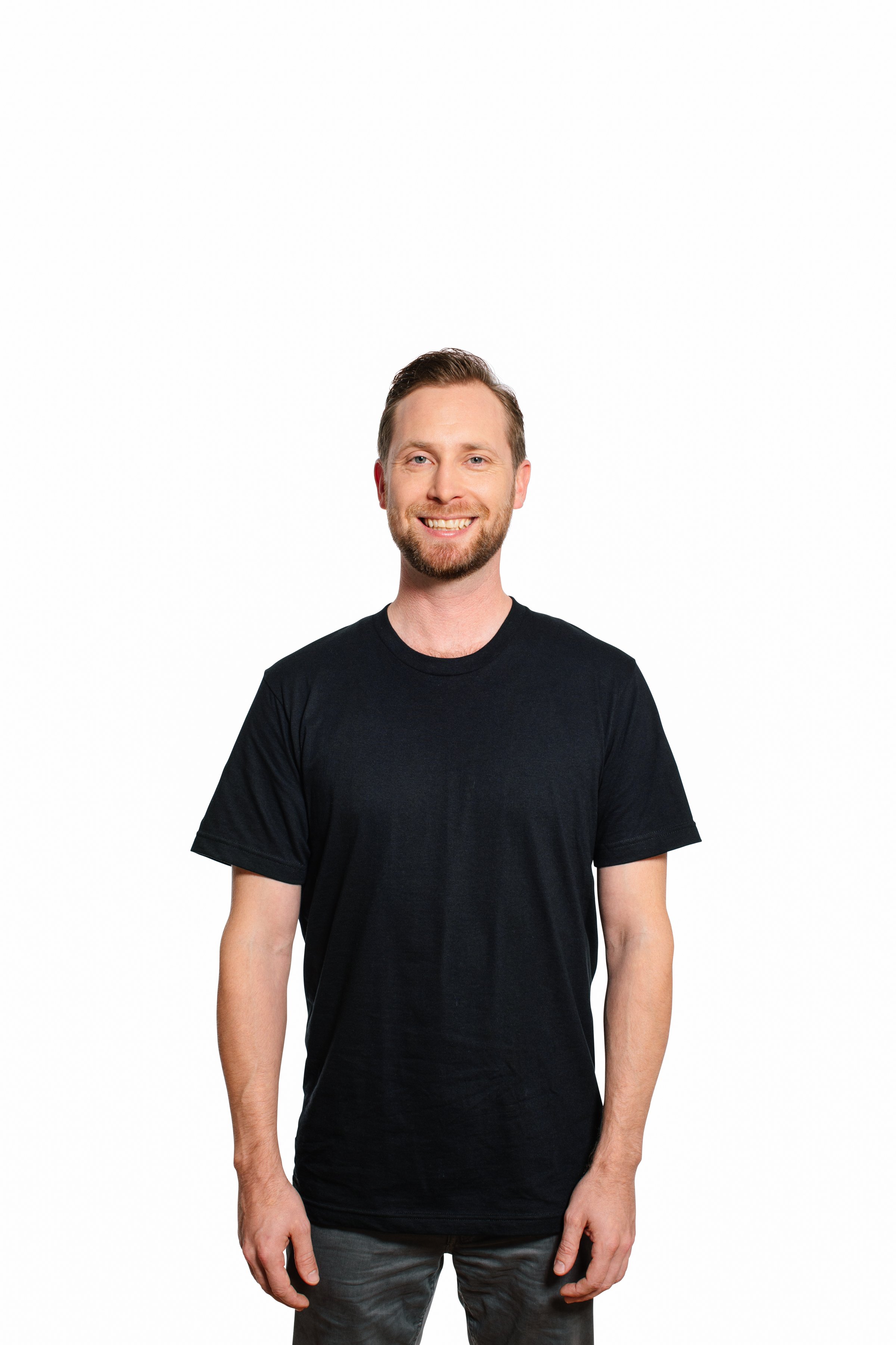 Smiling man with a beard wearing a black t-shirt and gray pants standing against a white background.