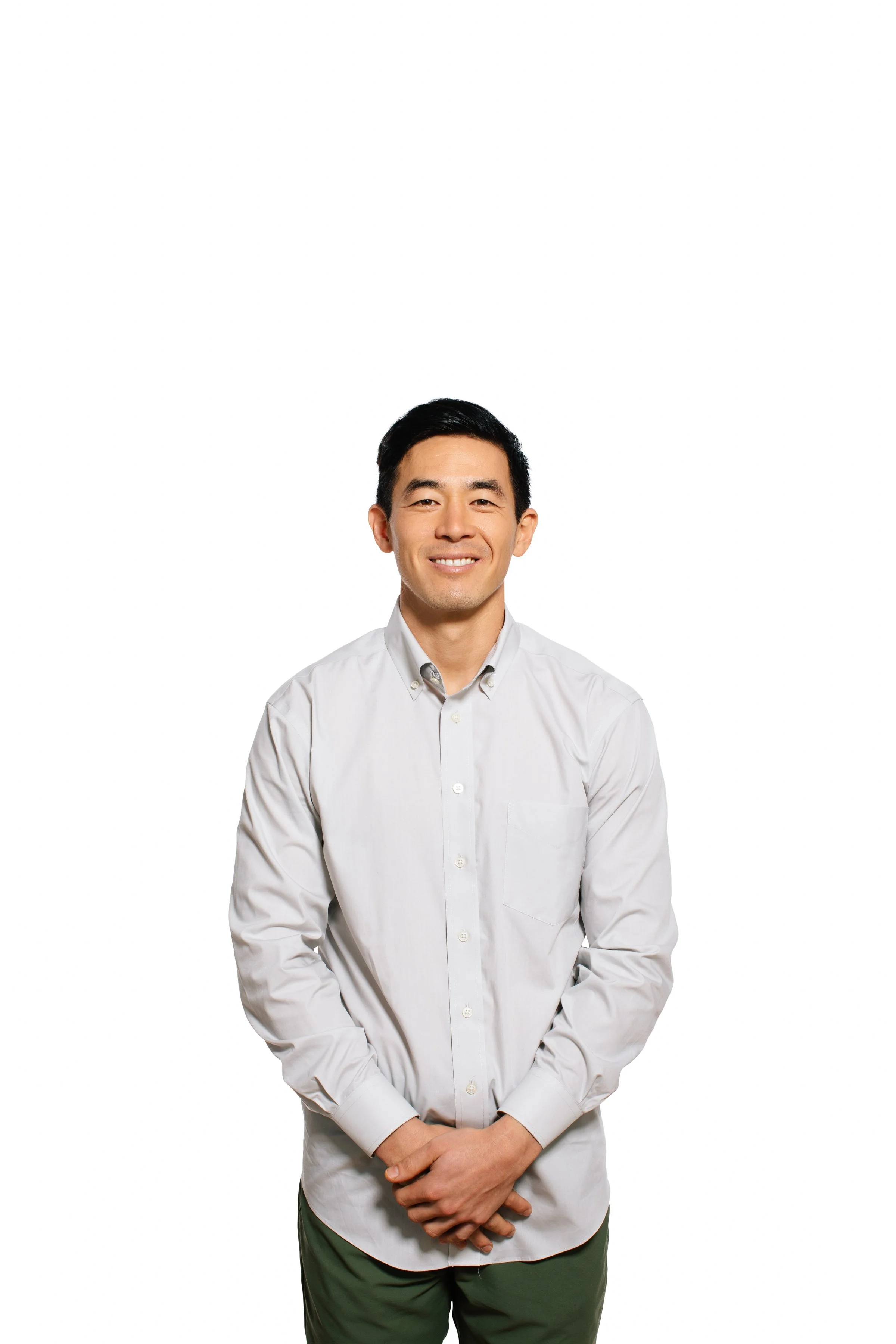 A man standing with his hands clasped in front of him, wearing a white button-up shirt and dark green pants, smiling against a plain white background.
