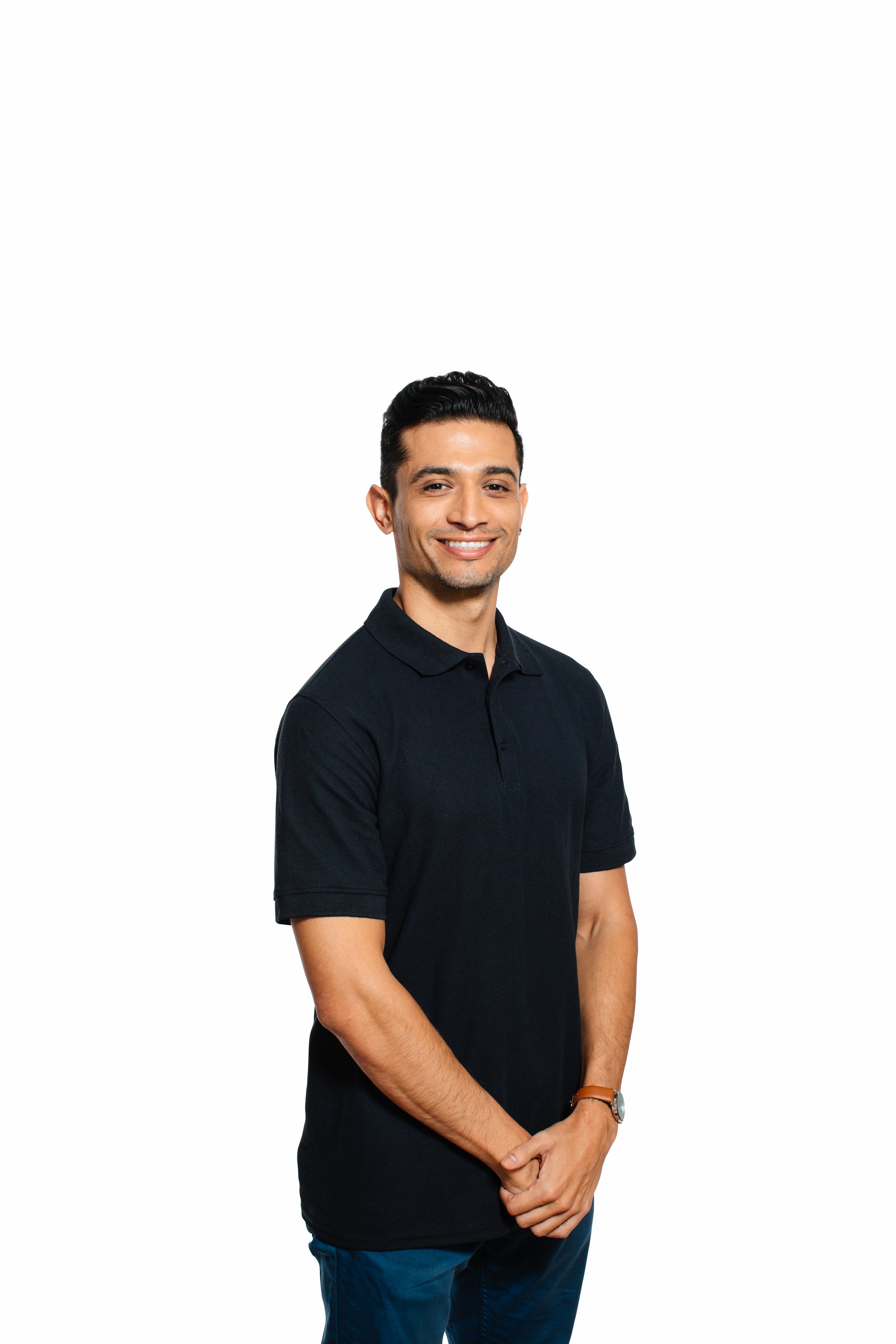 A young man with dark hair, smiling, wearing a dark short-sleeve collared shirt and blue pants, standing with his hands clasped in front of him against a white background.