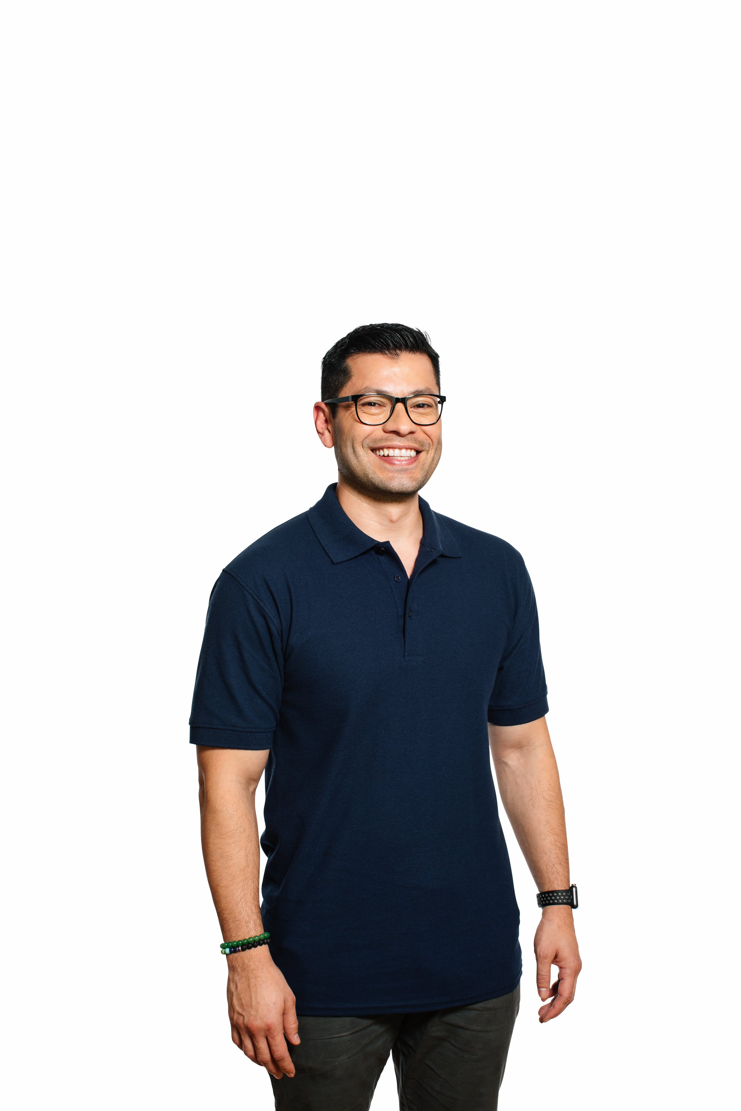 A man with short black hair, glasses, and light skin, smiling, wearing a navy blue polo shirt, dark pants, and a watch and bracelets on his left wrist, standing against a plain white background.