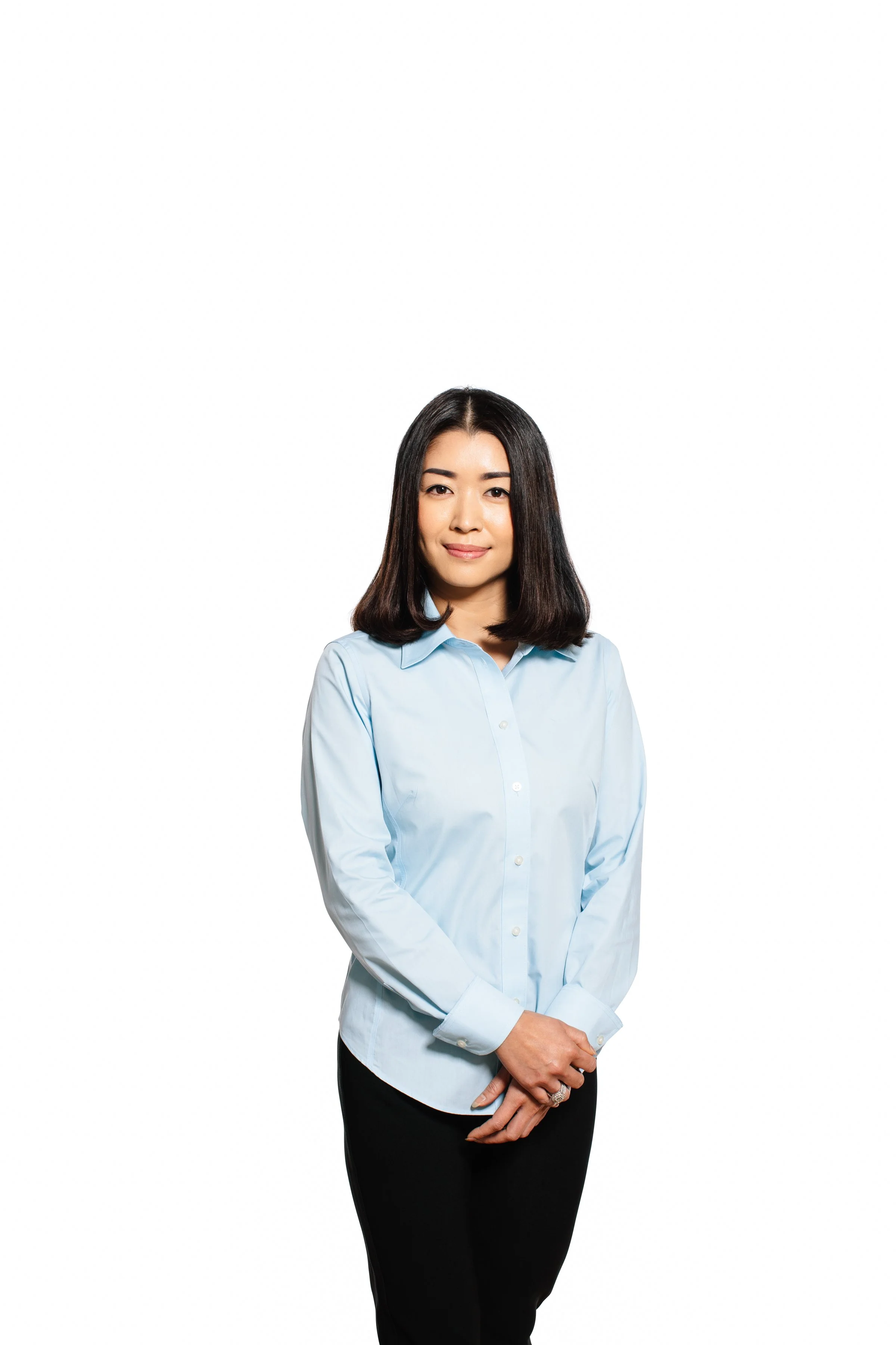 A woman with shoulder-length dark hair, wearing a light blue button-up shirt and black pants, standing with her hands clasped in front of her against a plain white background.