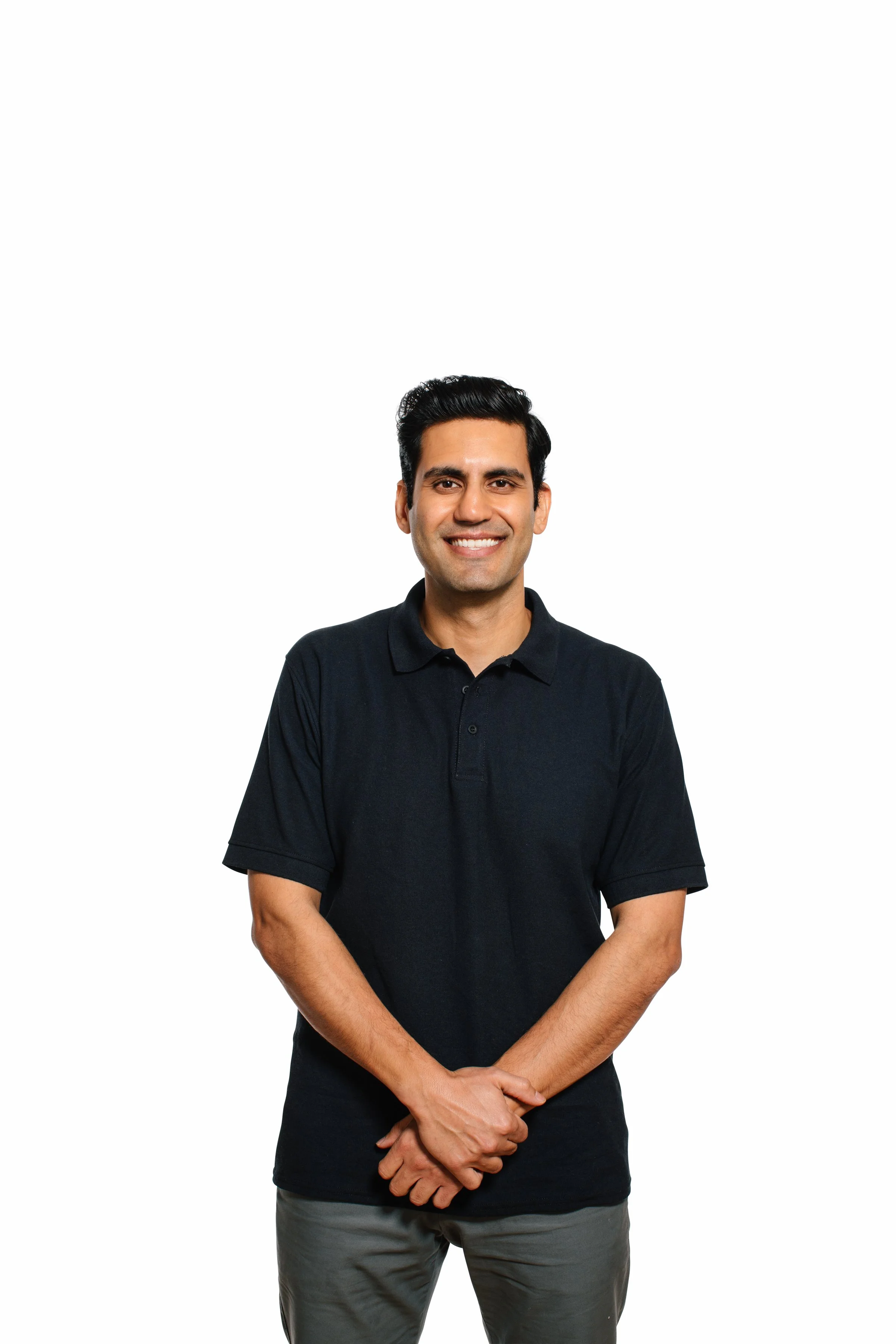A smiling man with dark hair wearing a black polo shirt and gray pants, standing against a white background.