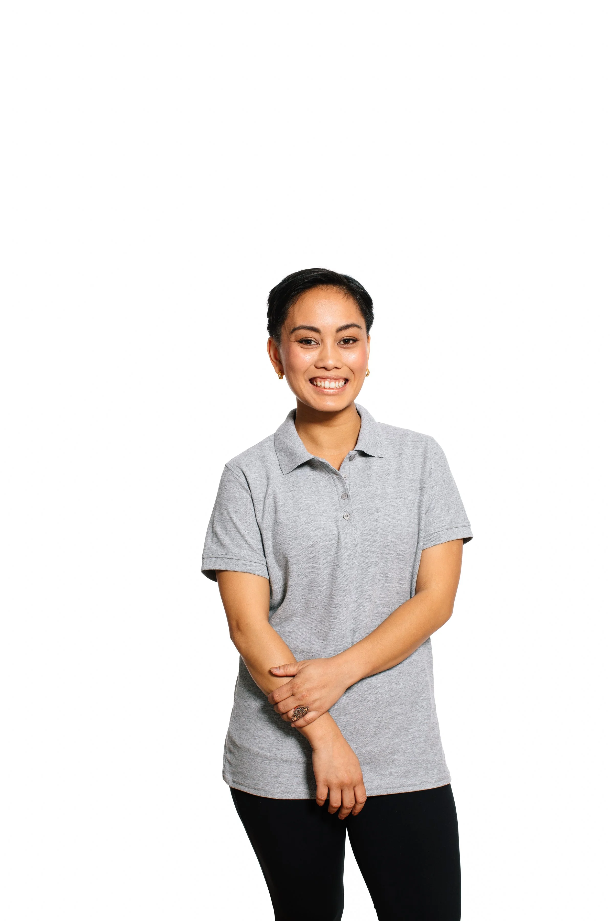 A smiling woman with short black hair, wearing a gray polo shirt and black pants, standing against a white background.