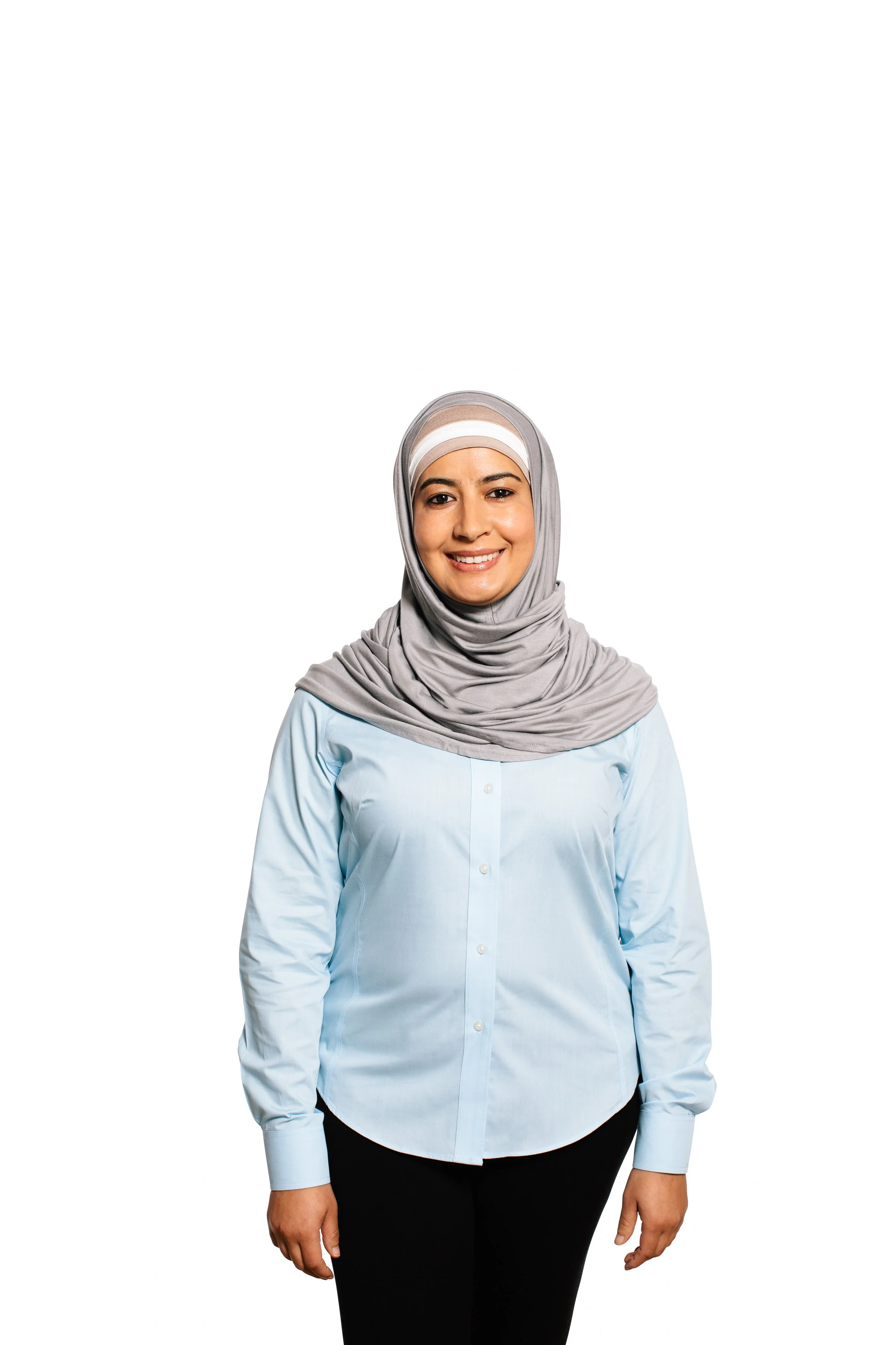 A woman wearing a light gray hijab, a light blue button-up shirt, and black pants standing against a white background.