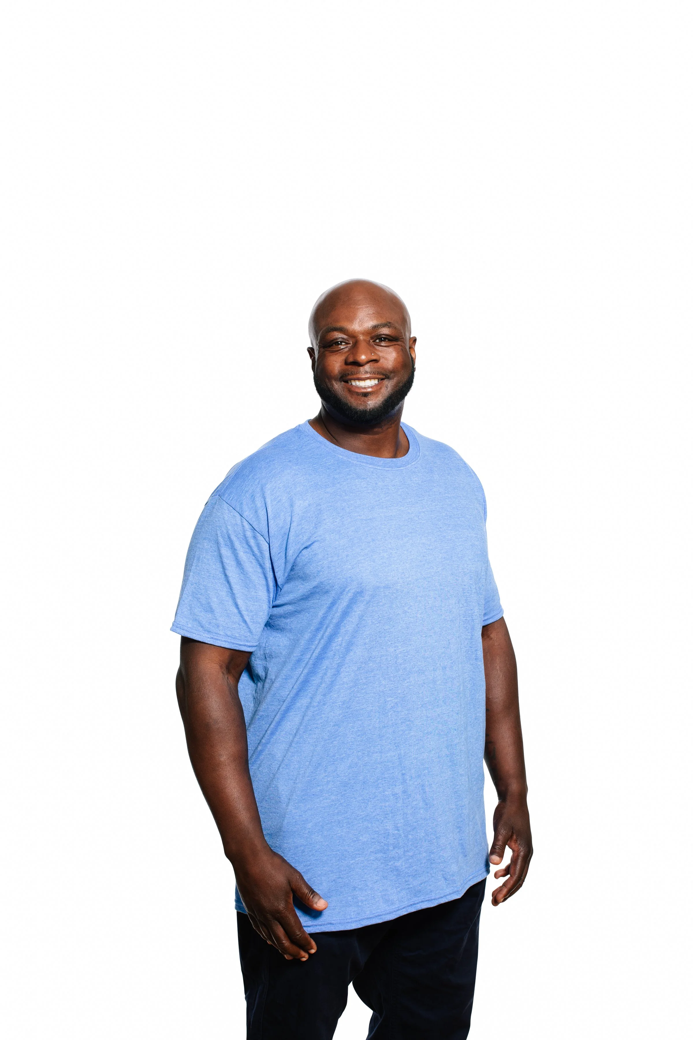 Smiling man with a shaved head and beard wearing a light blue T-shirt and black pants, standing against a plain white background.