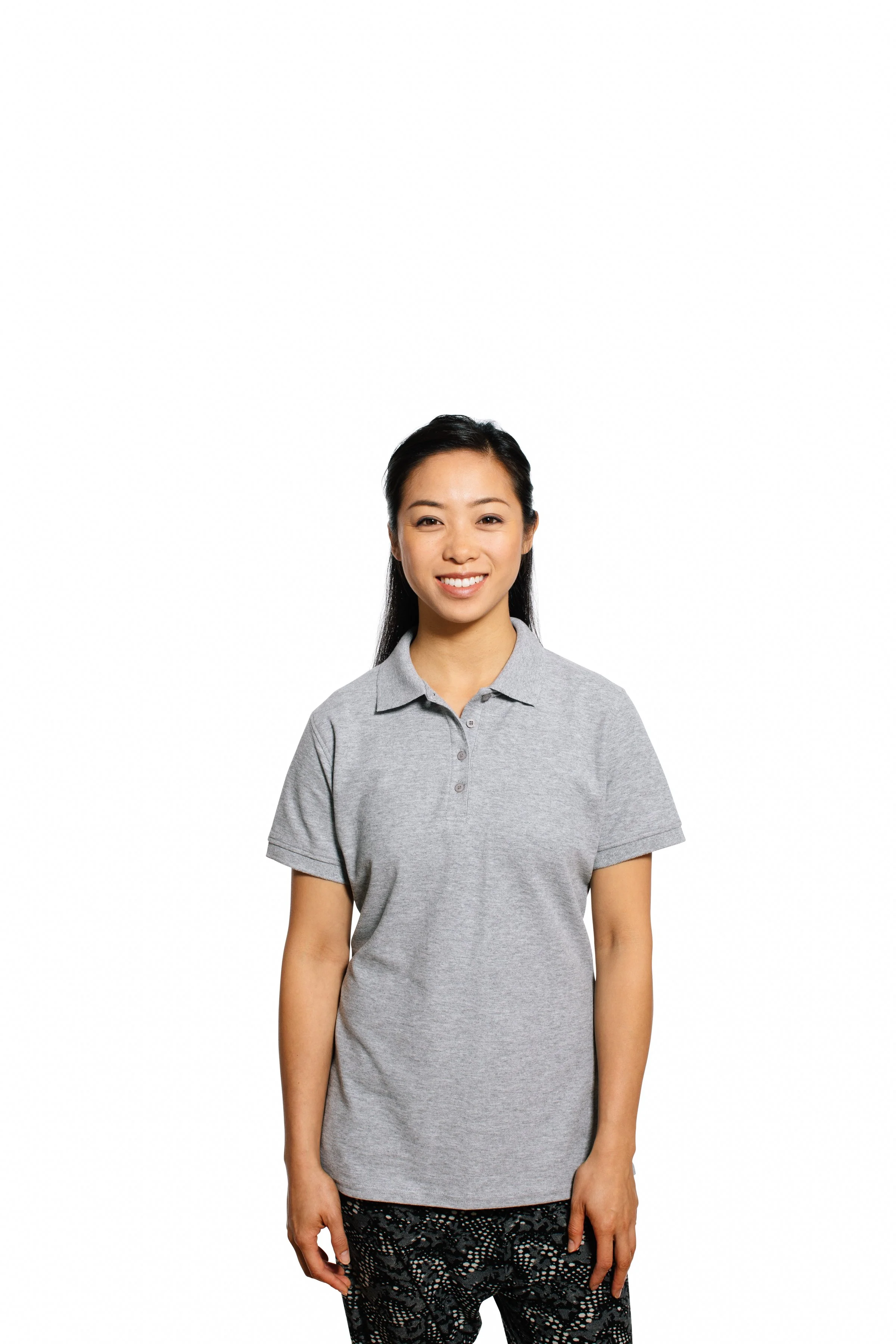 A young woman with long dark hair, smiling, wearing a gray polo shirt and patterned black pants, standing against a white background.