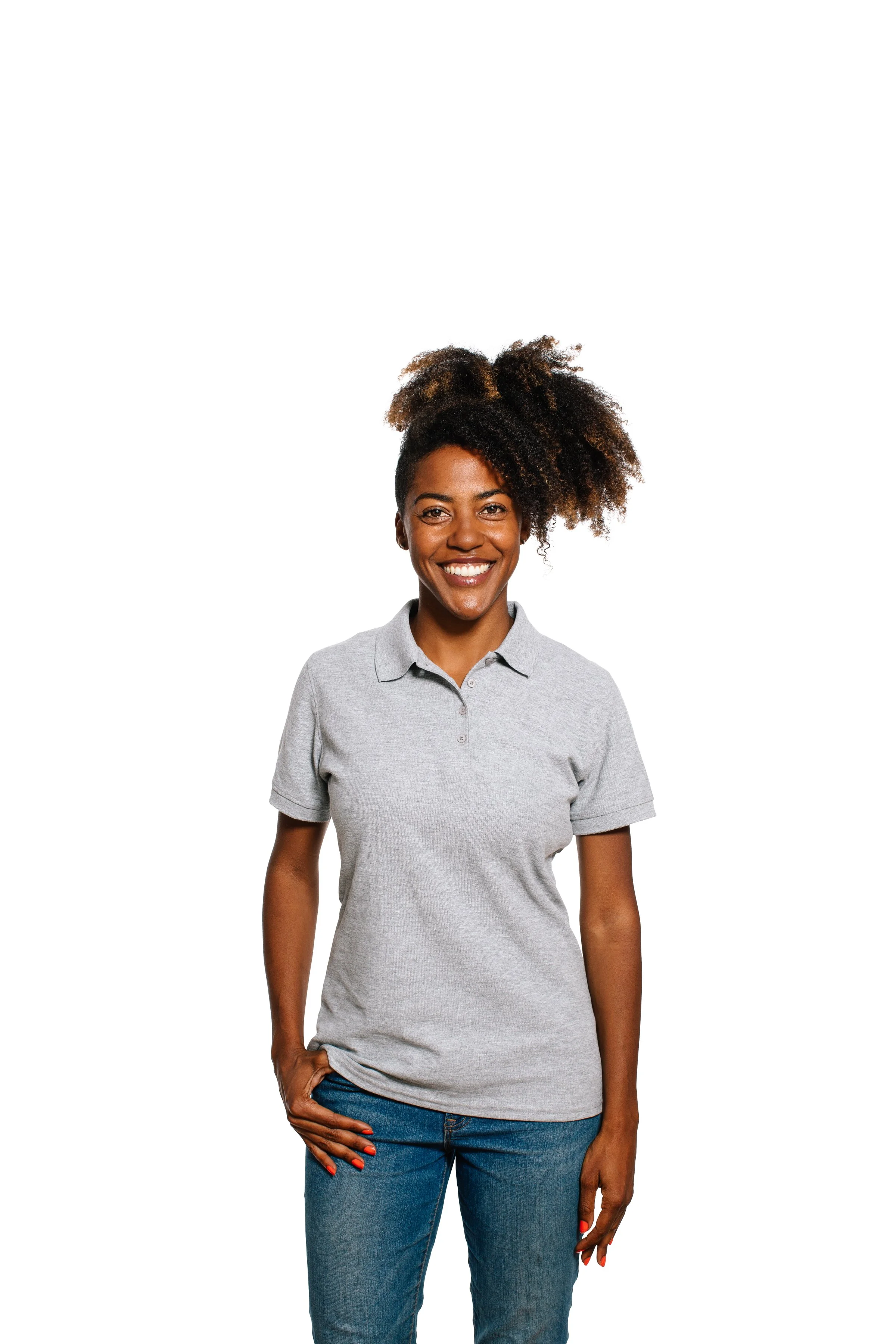 A woman with dark skin and curly hair styled in an updo, smiling, wearing a light gray polo shirt and blue jeans, standing against a plain white background.
