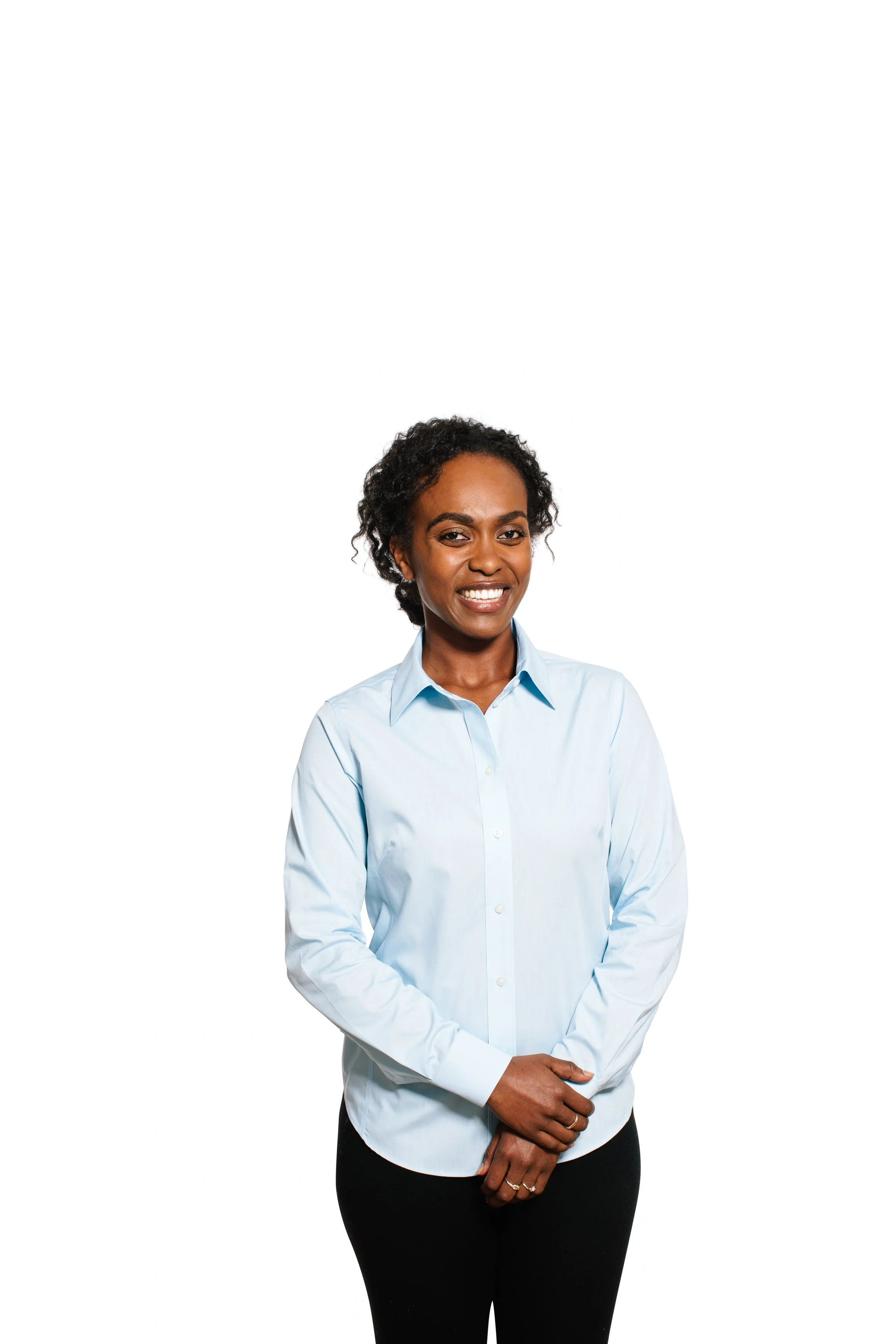 A woman with short curly hair wearing a light blue button-up shirt and black pants standing against a plain white background.