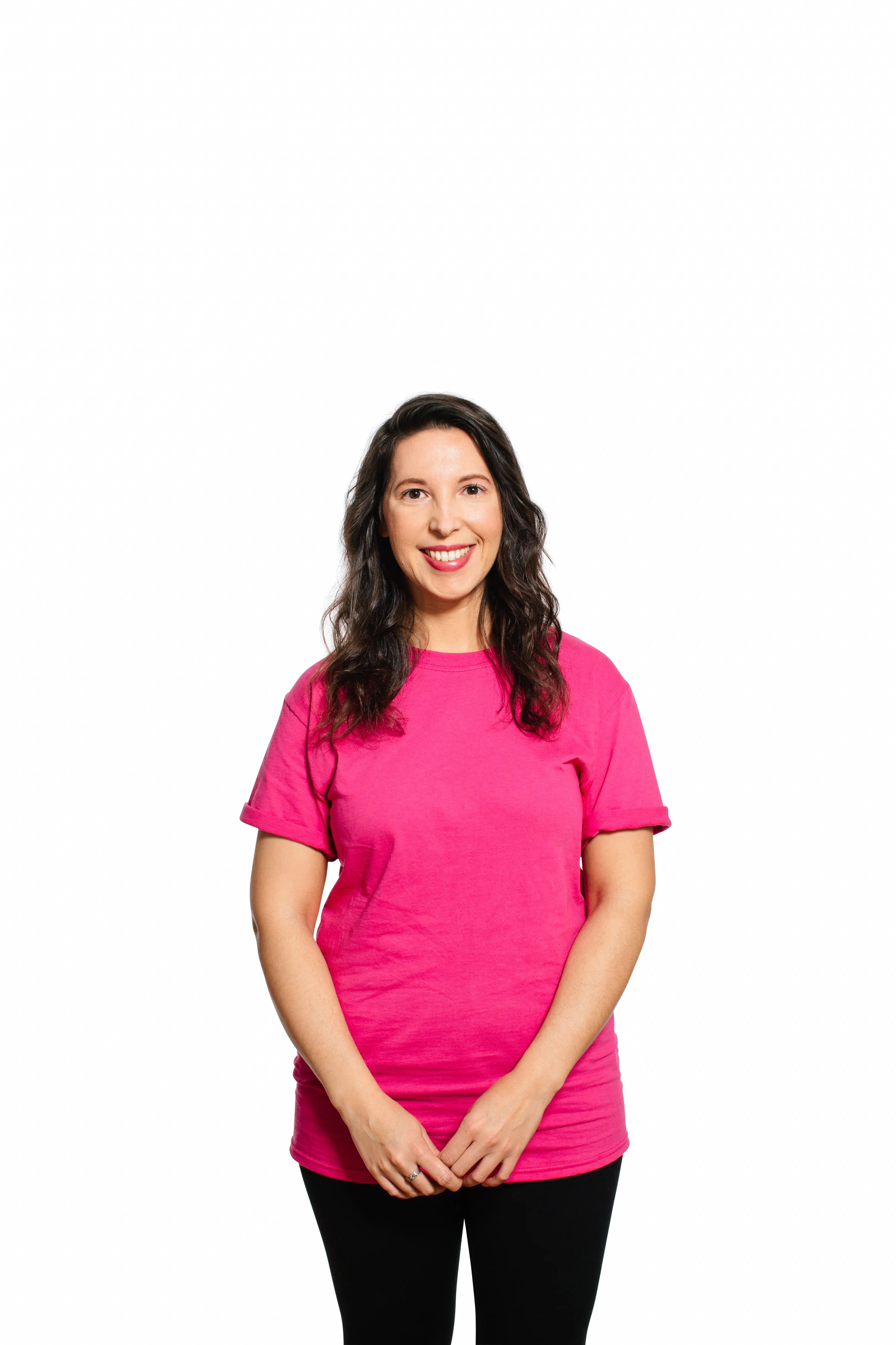 A woman with long dark hair wearing a bright pink short-sleeved T-shirt, standing against a plain white background. She is smiling and has her hands clasped in front of her.