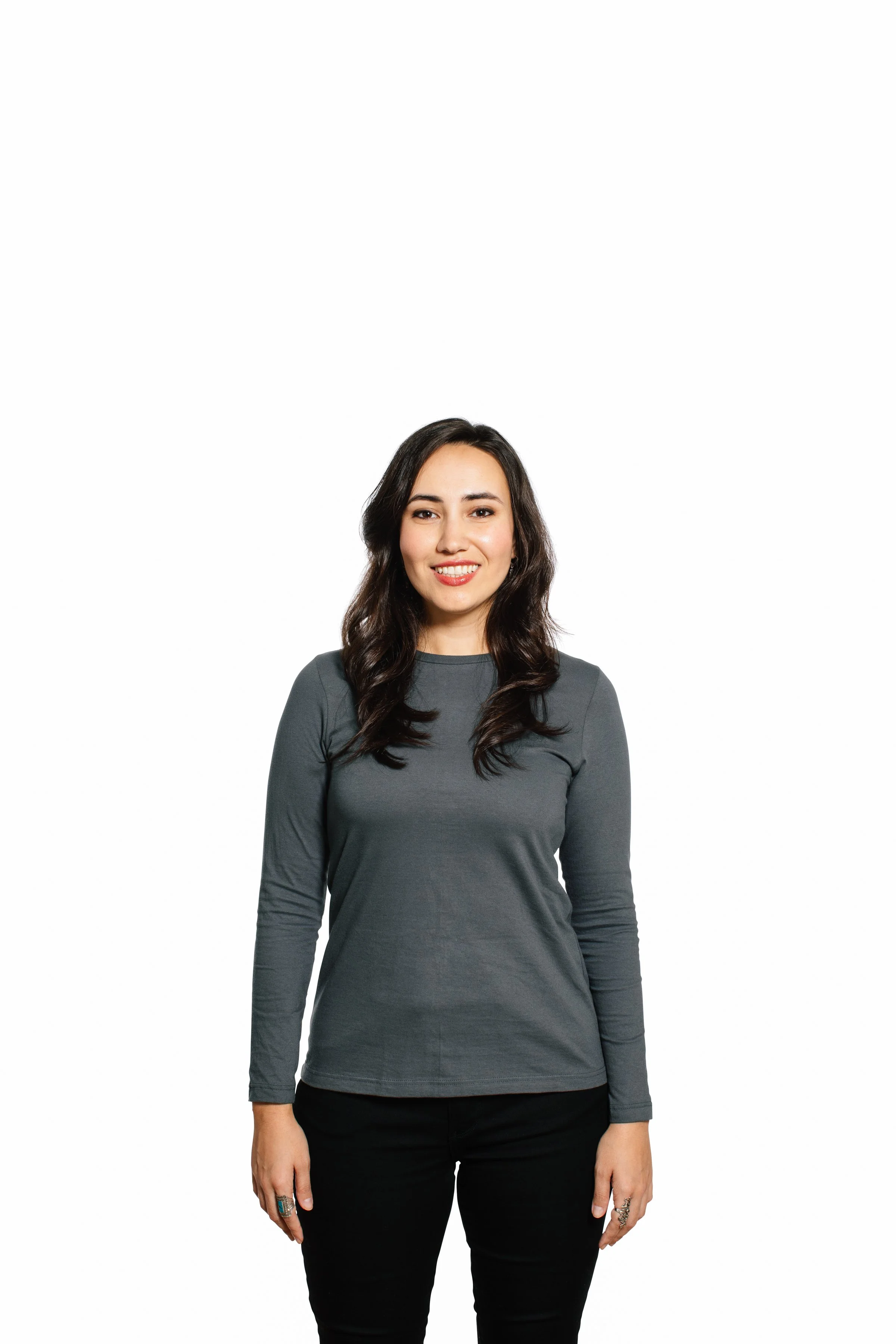 A woman standing against a white background, smiling, wearing a long-sleeved gray shirt and black pants.