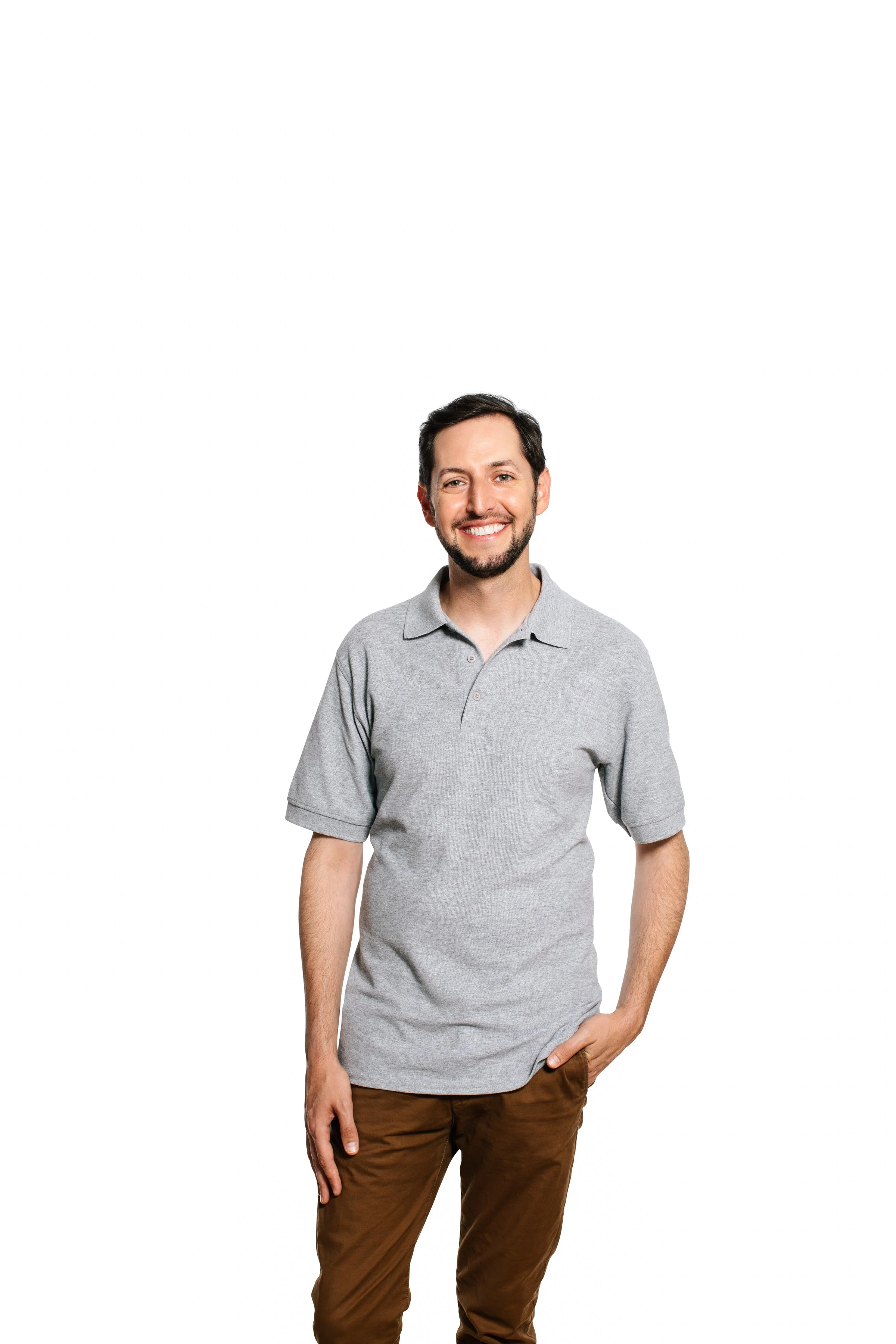 A man with dark hair, a beard, and a smile wearing a light gray polo shirt and brown pants, standing against a white background.