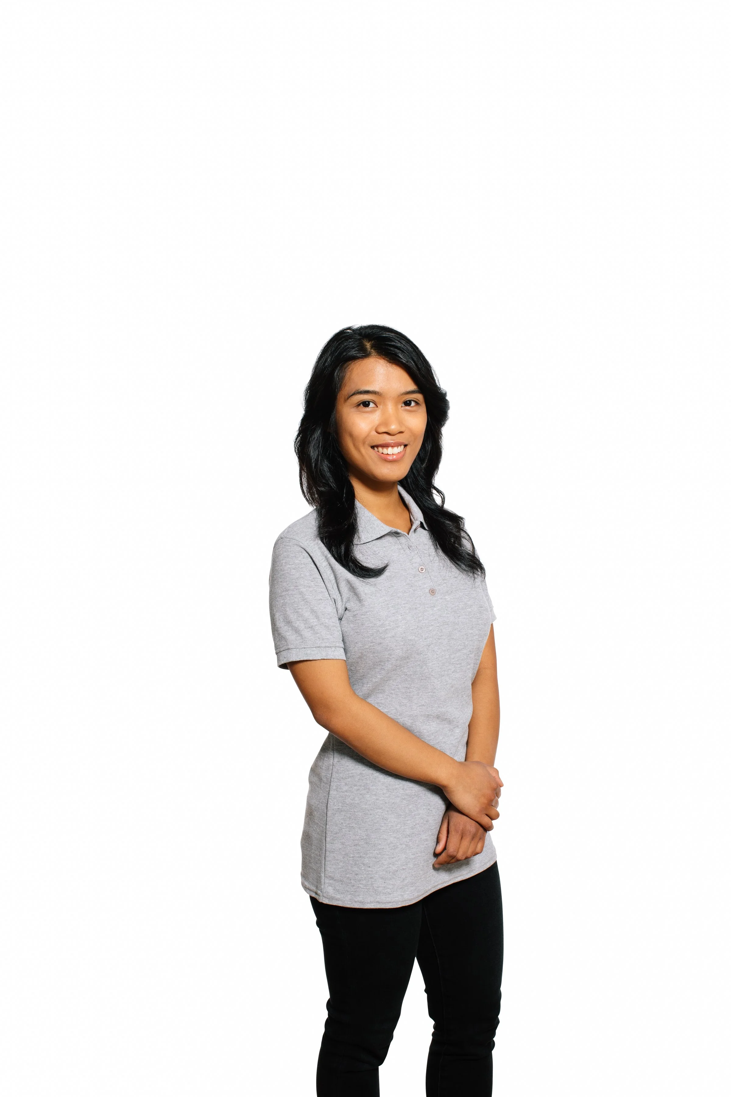 A woman with black hair wearing a gray polo shirt and black pants, standing with her hands clasped in front of her against a white background.