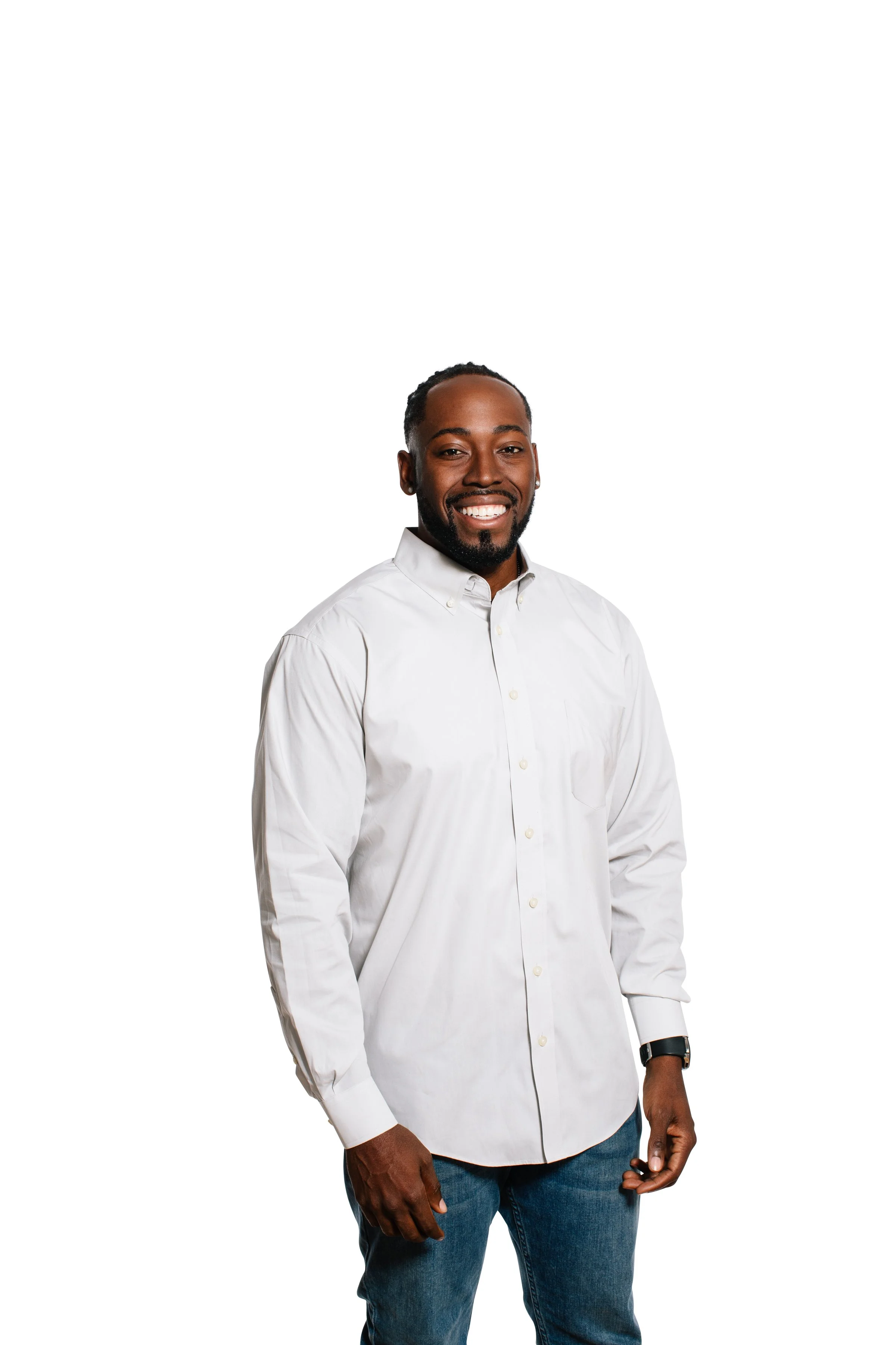 A smiling man with dark skin, wearing a white button-up shirt and blue jeans, standing against a plain white background.