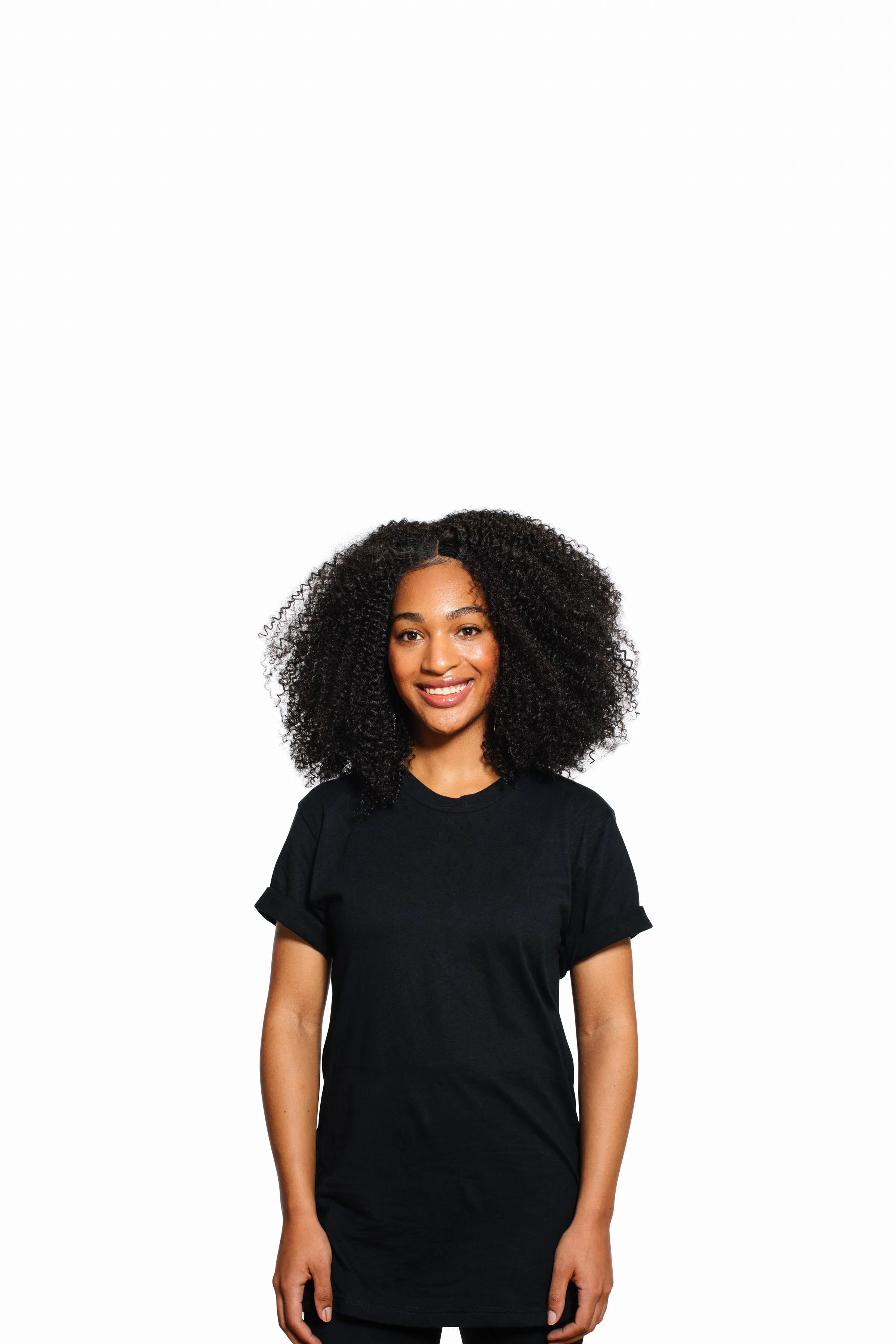 A young woman with curly black hair wearing a black t-shirt, smiling and standing against a plain white background.