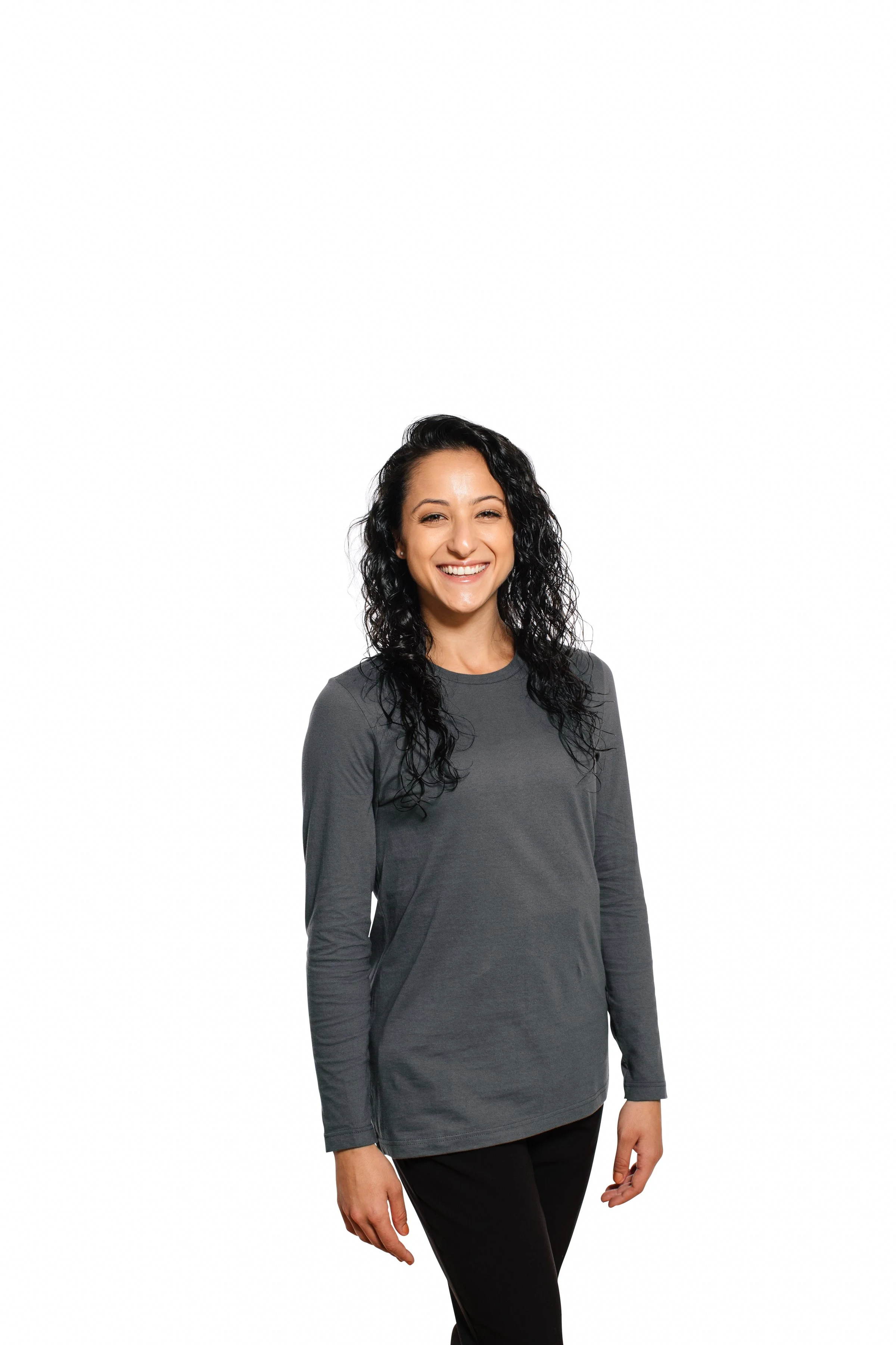 A woman with curly black hair smiling, wearing a long-sleeved dark gray shirt and black pants, standing against a white background.