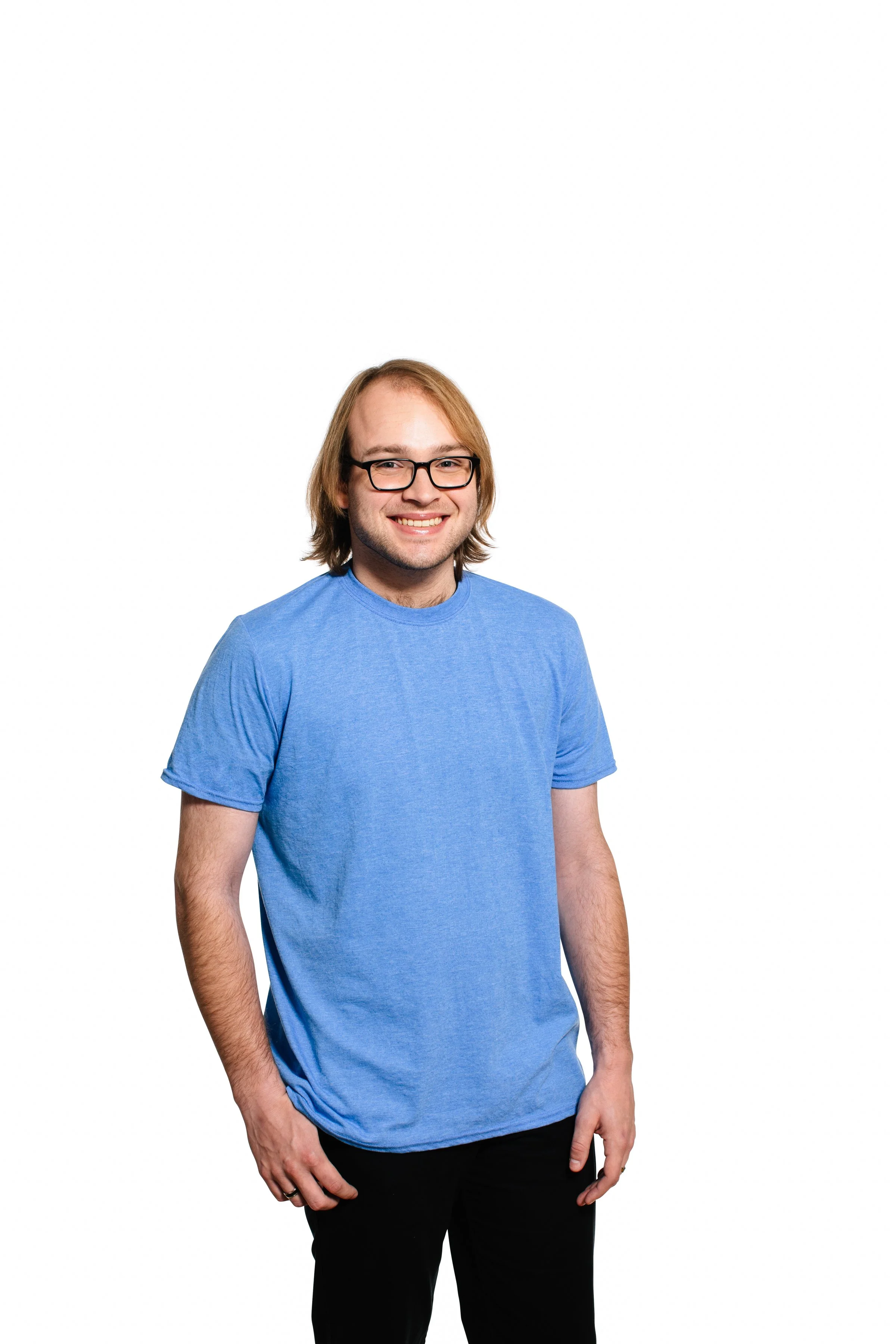 A young man with glasses and shoulder-length blond hair, smiling, wearing a blue t-shirt and black pants, standing against a plain white background.