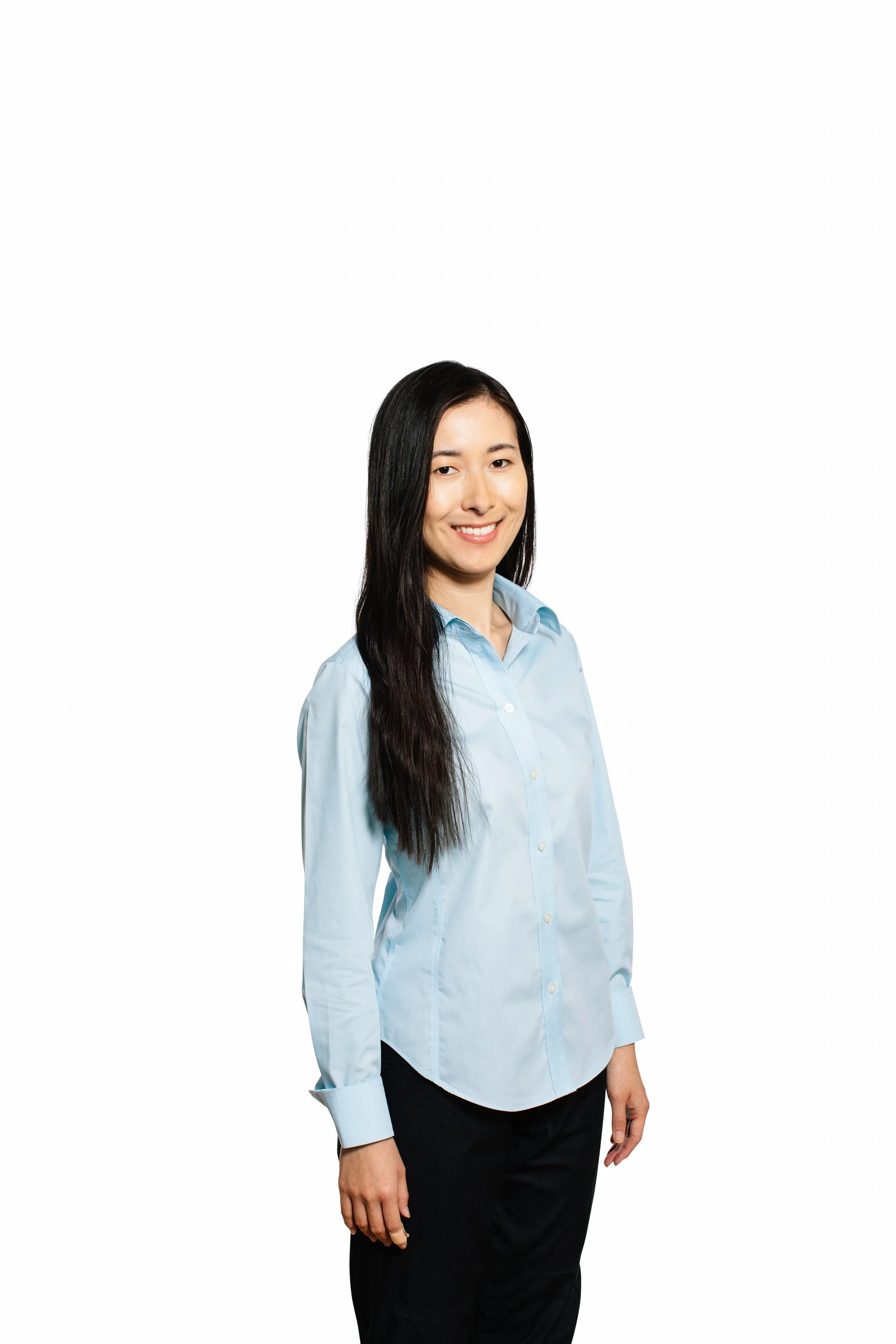A woman with long dark hair smiling, wearing a light blue button-up shirt and black pants, standing against a white background.
