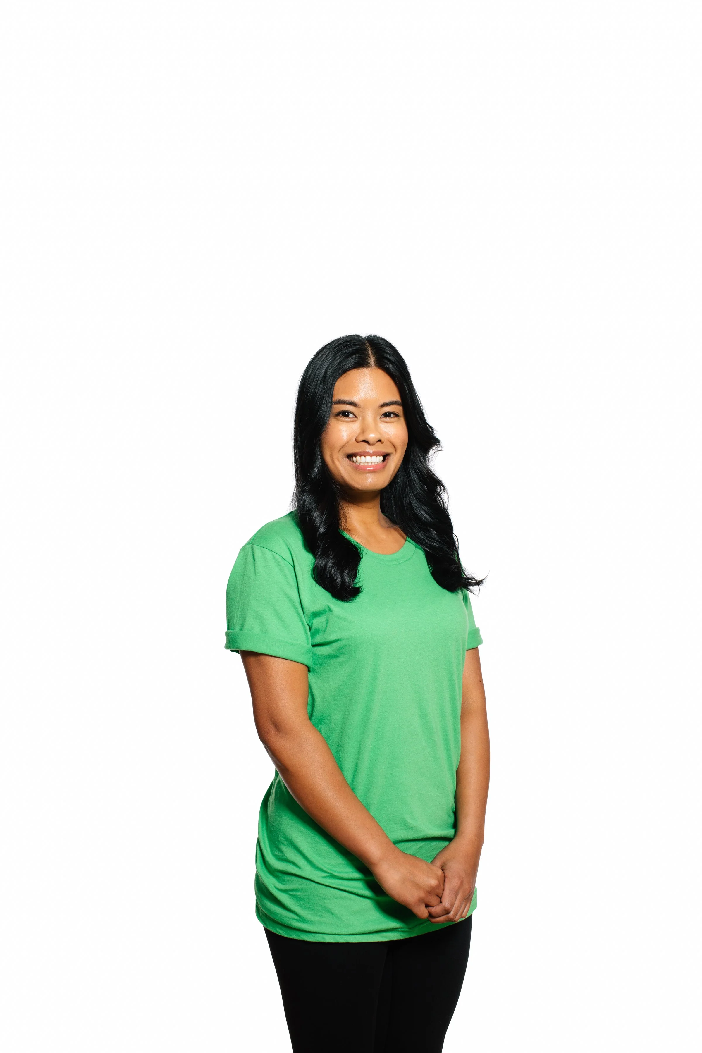 A woman with long black wavy hair smiling, wearing a bright green t-shirt and black pants, standing with her hands clasped in front of her against a plain white background.