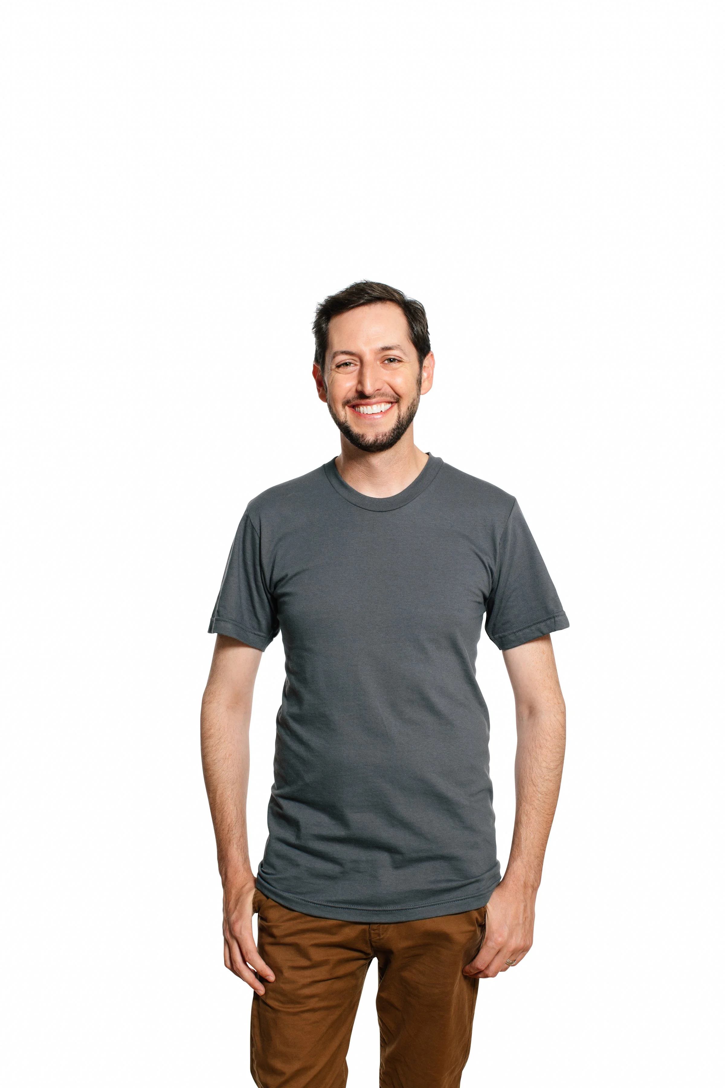 A young man with short dark hair and a beard, smiling and wearing a dark gray t-shirt and brown pants, standing against a plain white background.