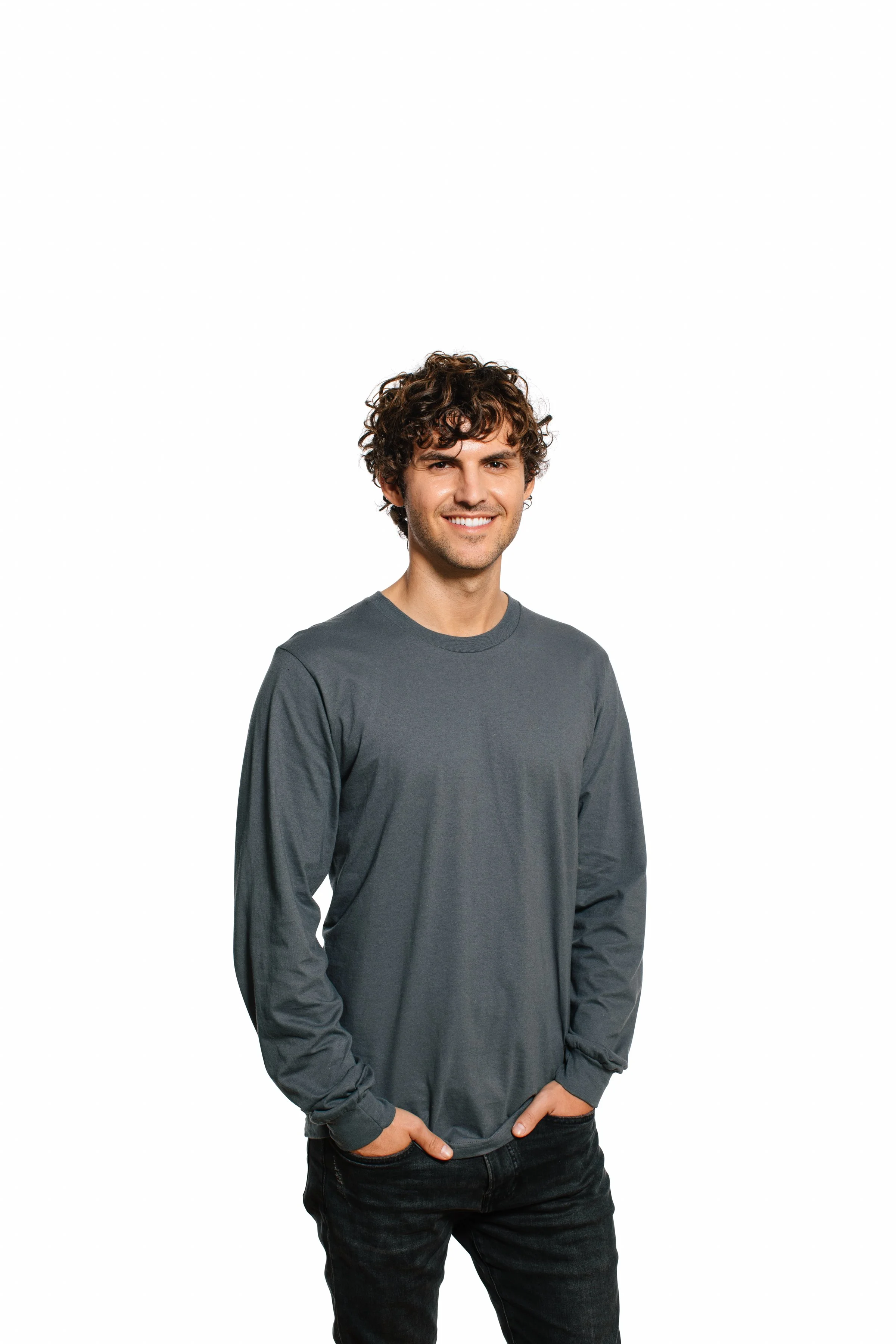 A young man with curly brown hair smiling, wearing a long-sleeved dark gray shirt and black jeans, standing against a plain white background.