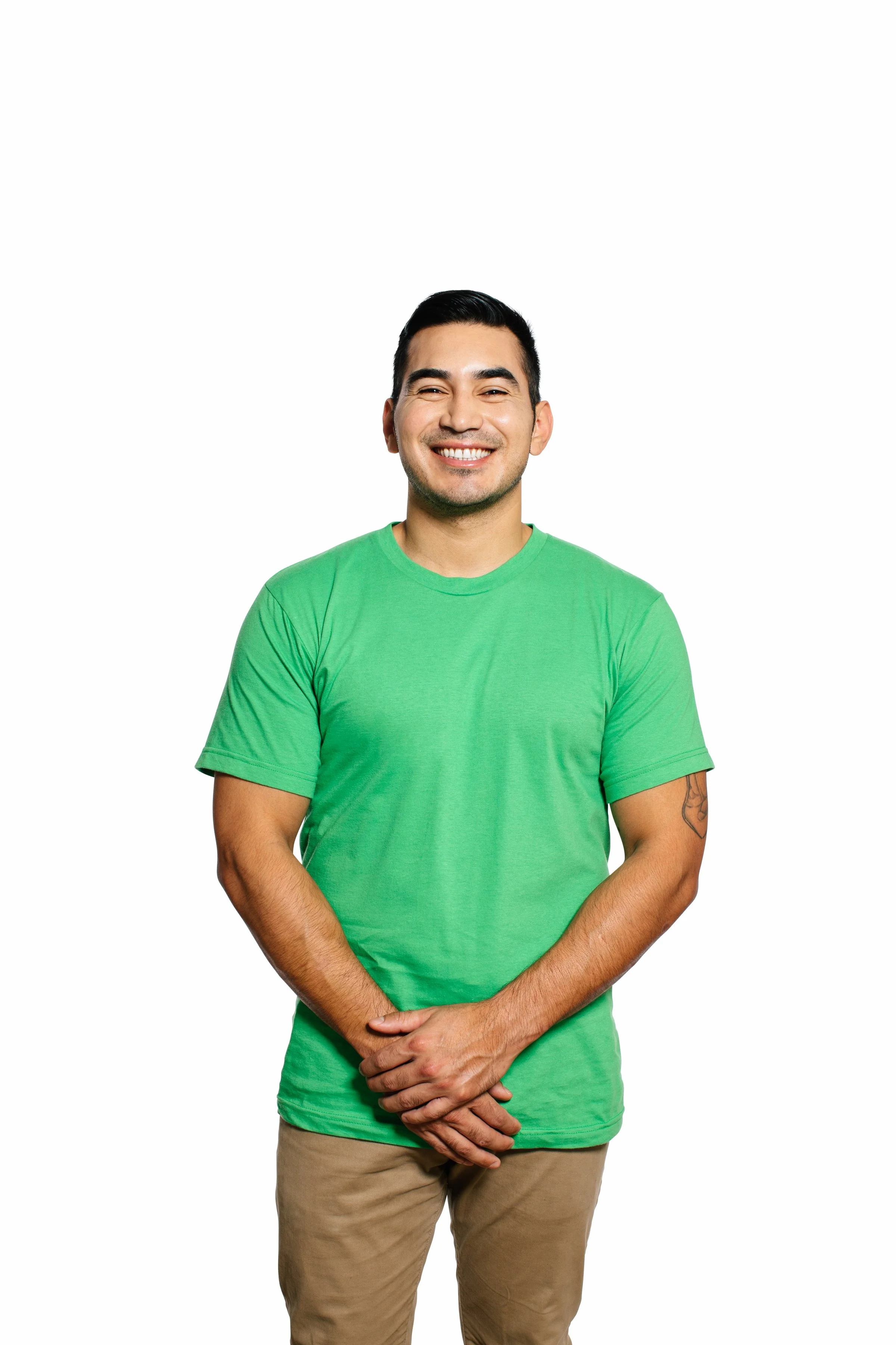 A smiling man with dark hair and a short beard, wearing a bright green t-shirt and khaki pants, standing against a plain white background with his hands clasped in front of him.
