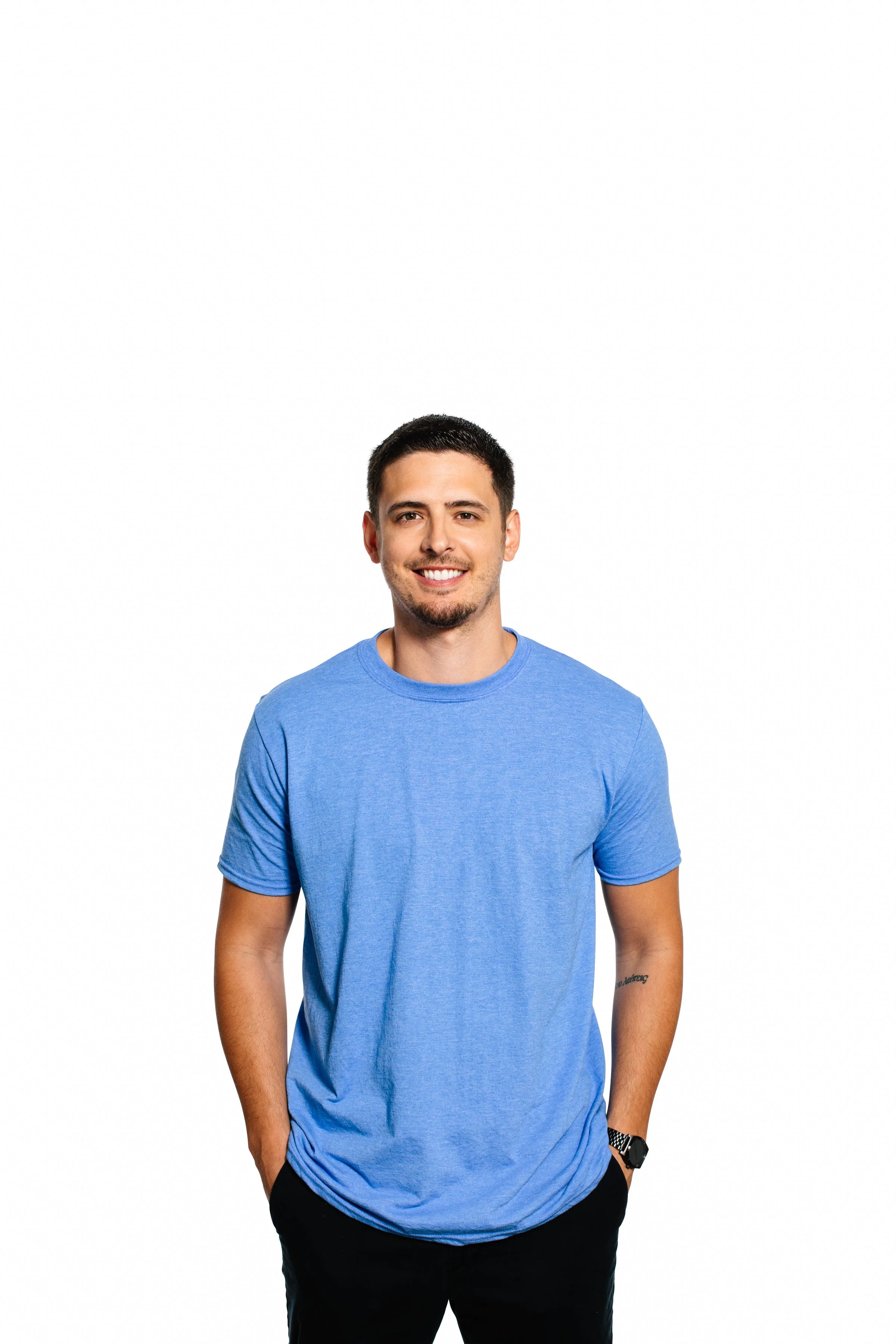 A young man with short dark hair, a trimmed beard, and a friendly smile, wearing a blue T-shirt and standing with his hands in his pockets against a plain white background.
