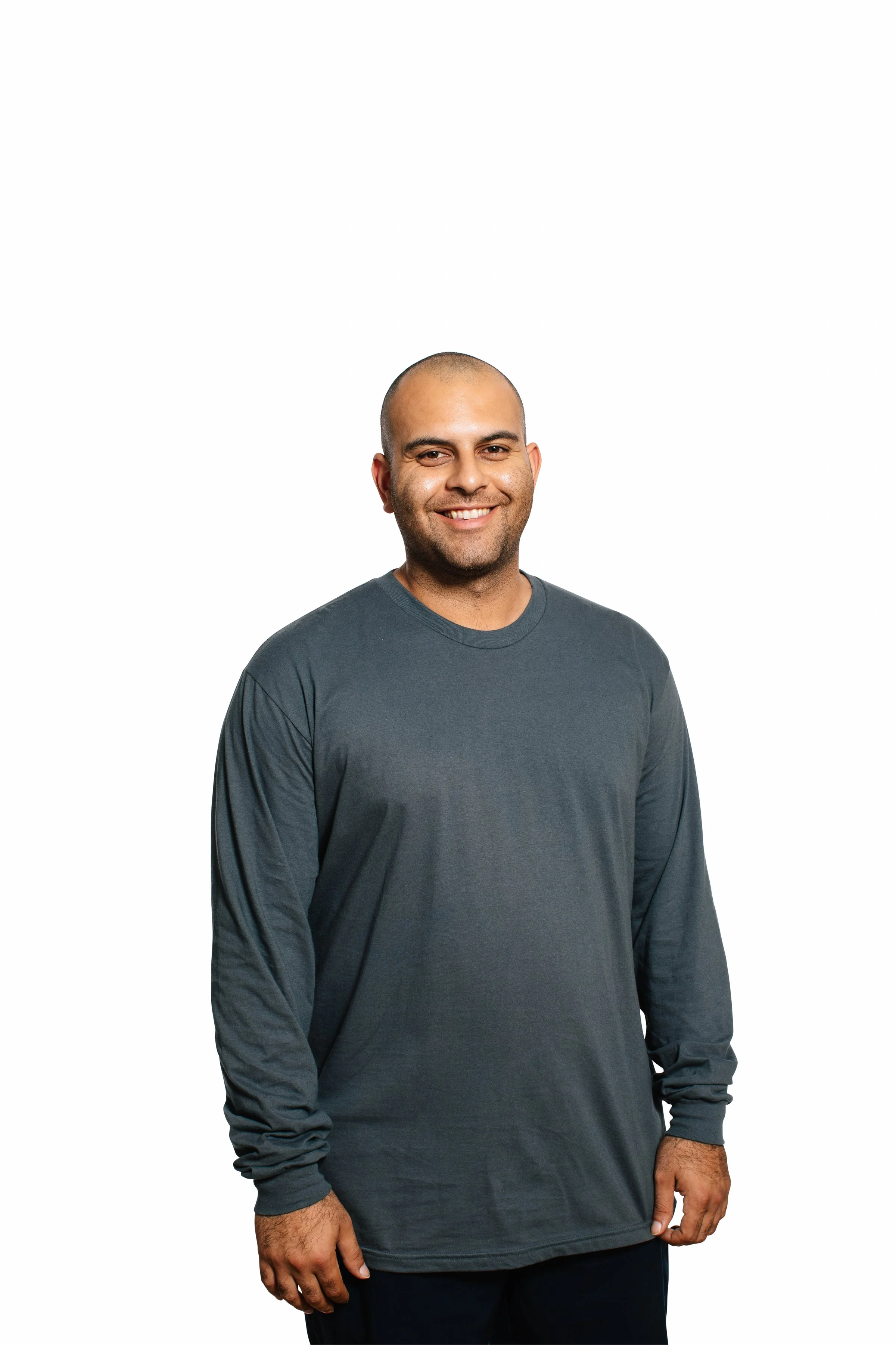 A smiling man with a shaved head wearing a long-sleeve dark gray shirt and dark pants, standing against a plain white background.