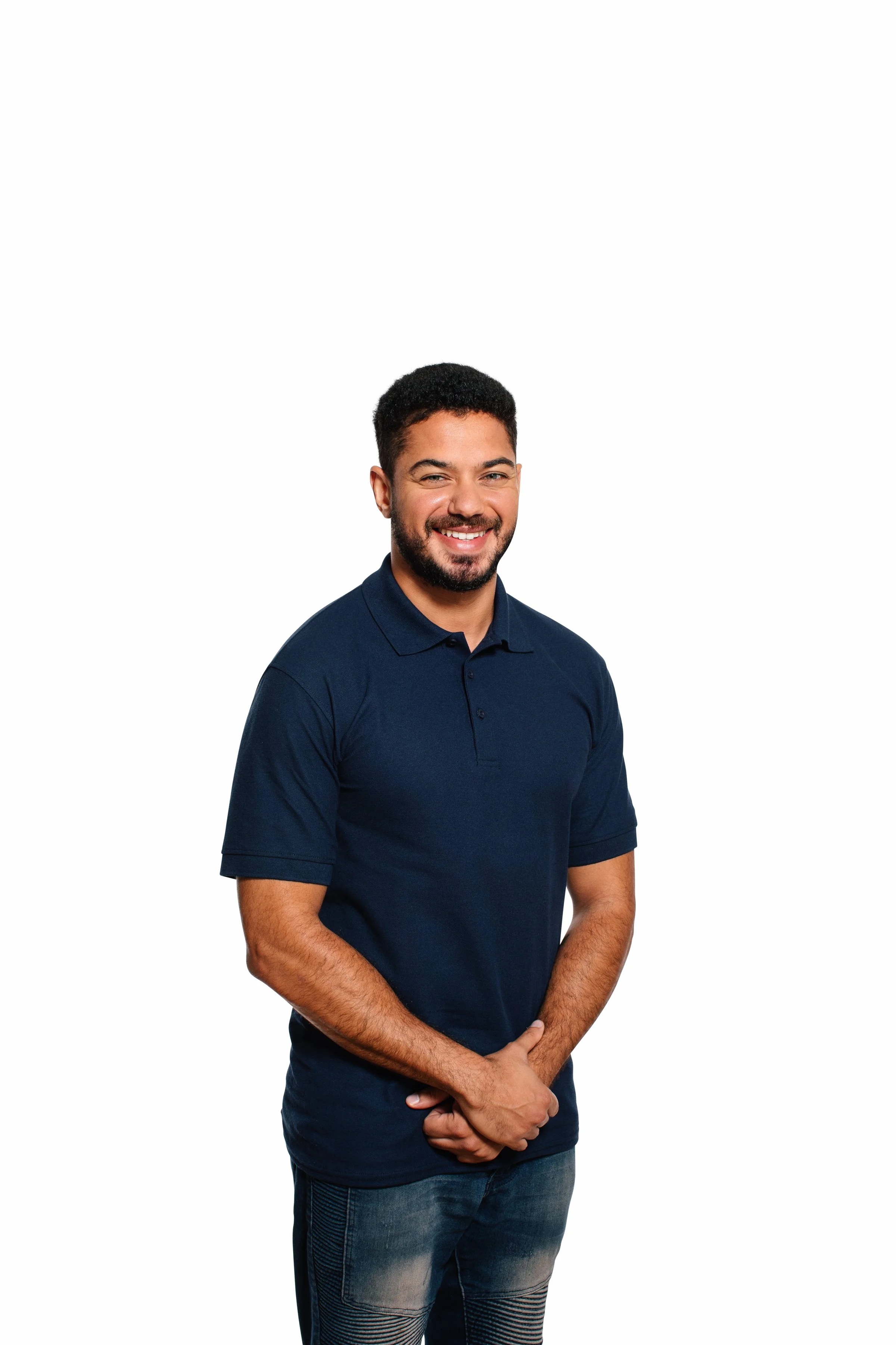 A man with dark hair and a beard wearing a navy blue polo shirt and jeans, standing with his hands clasped, smiling, against a white background.