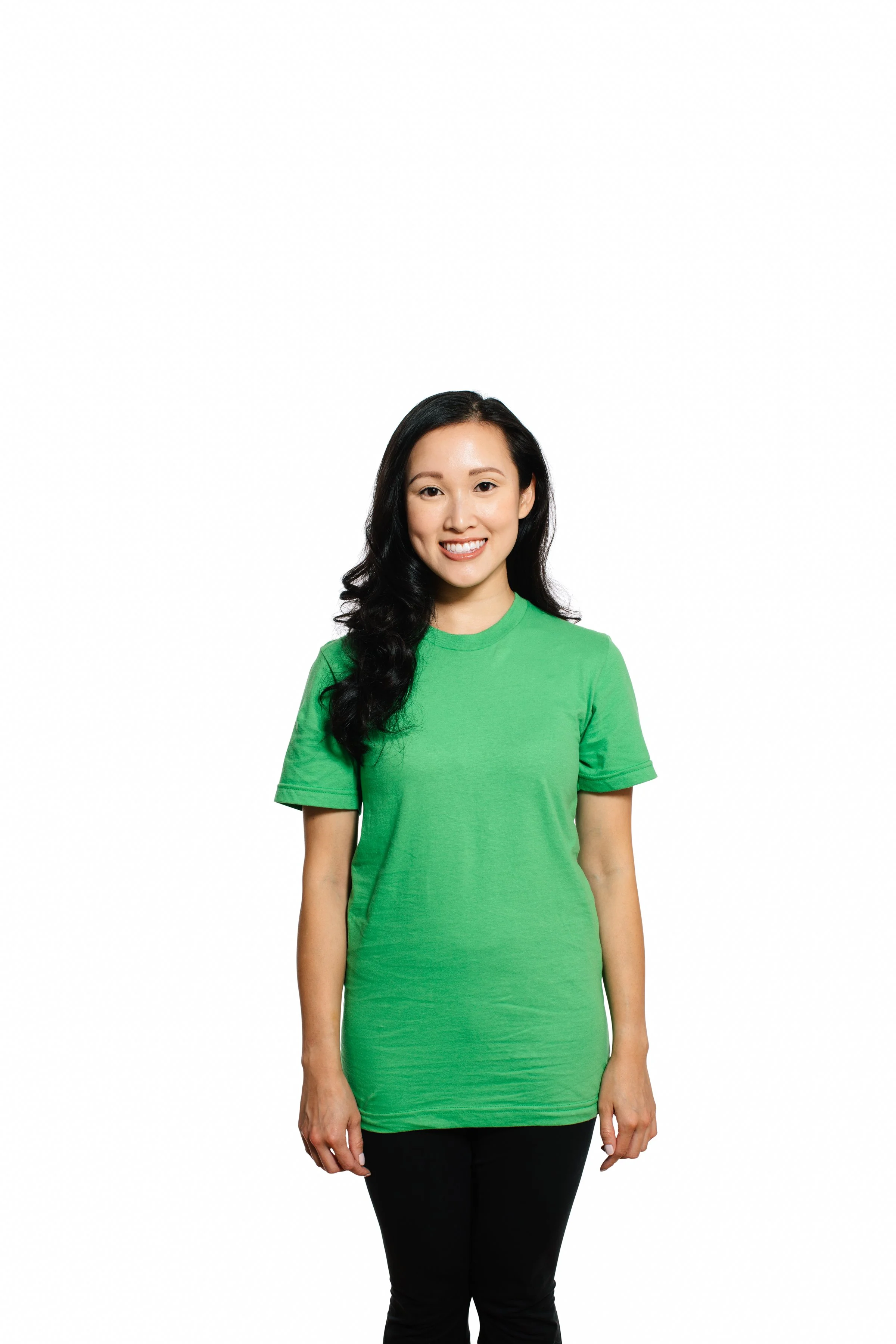 A woman with long black hair, smiling, wearing a green t-shirt and black pants, standing against a plain white background.