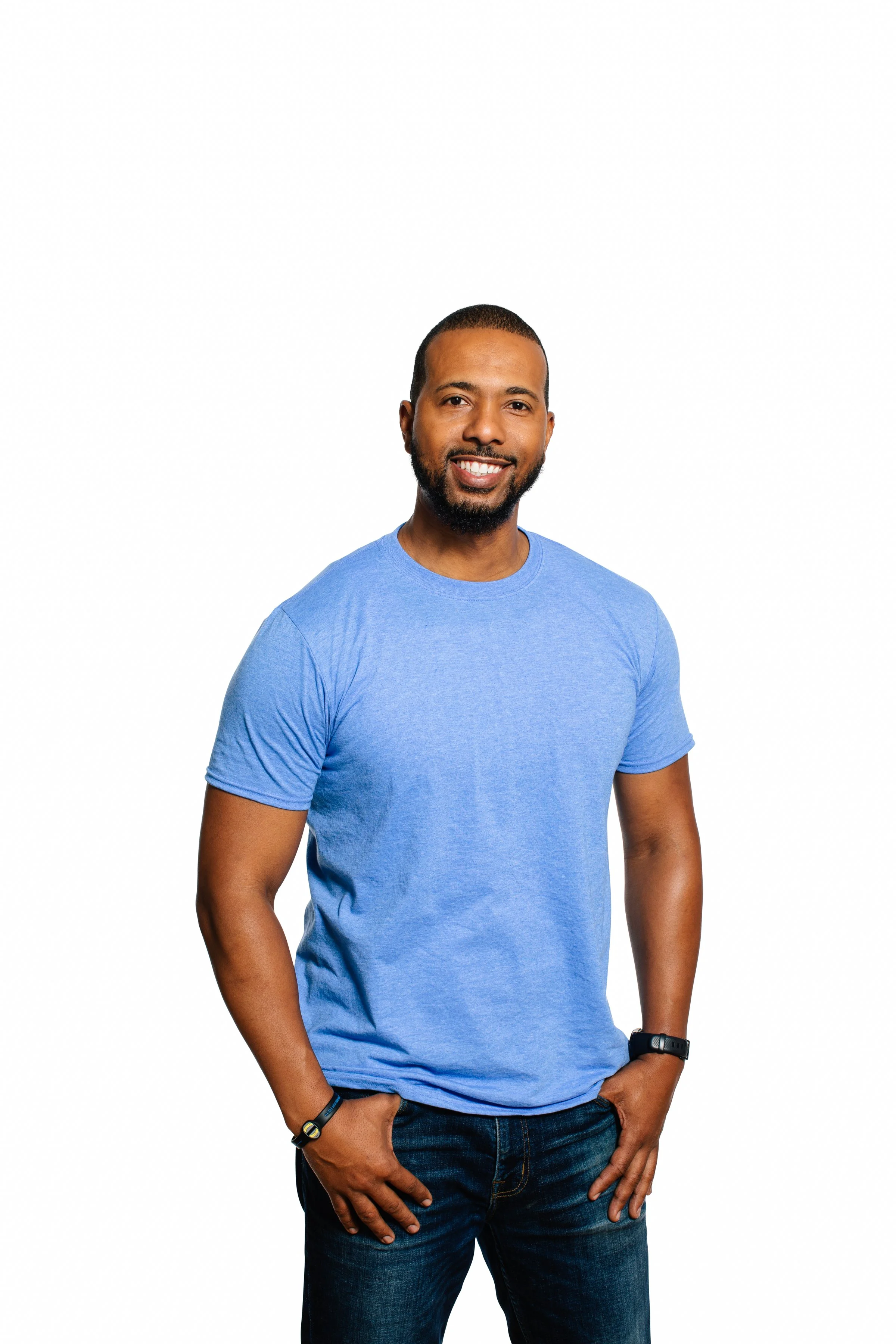A smiling man with short hair, a beard, wearing a blue t-shirt and jeans, standing with one hand in his pocket against a white background.