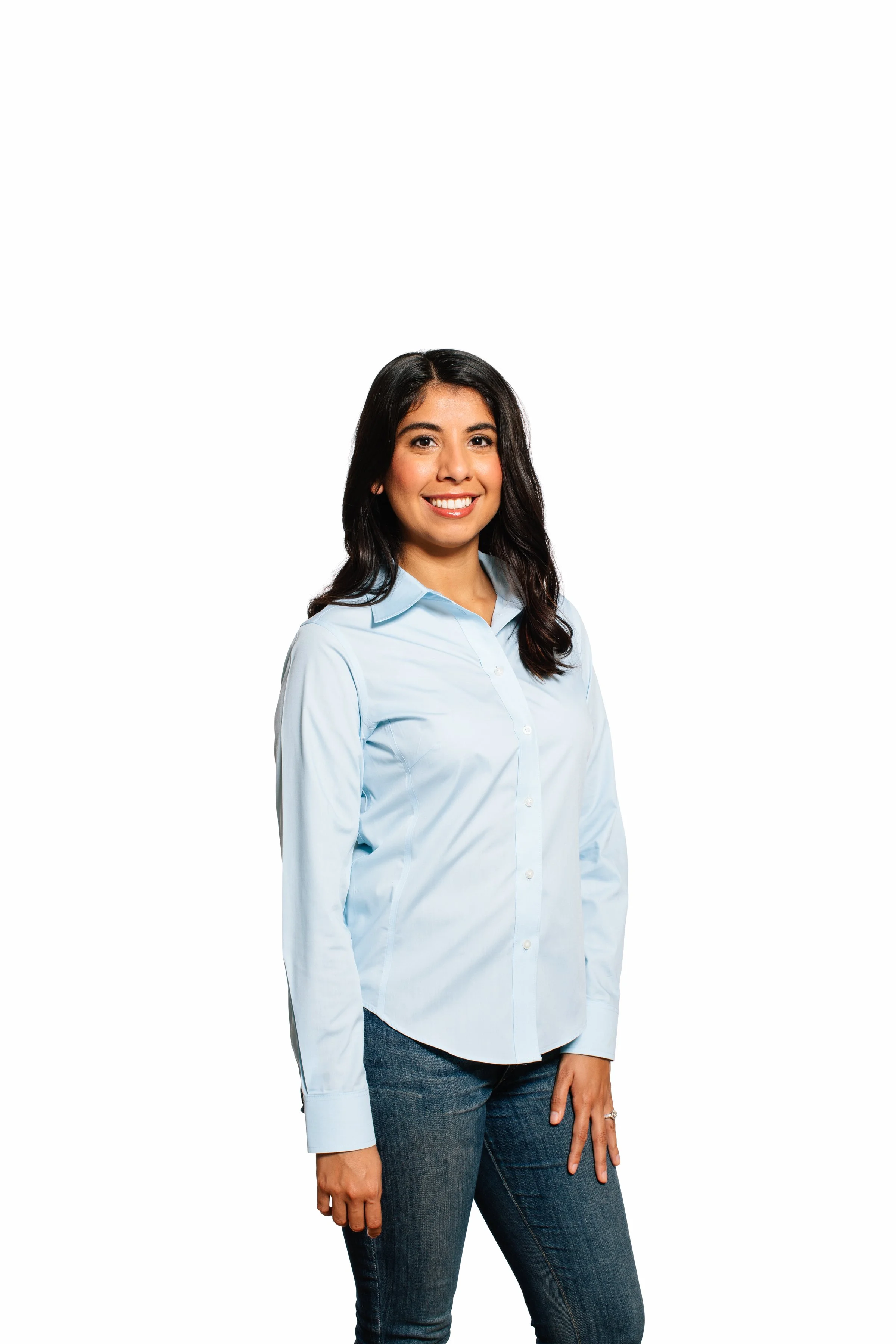 A portrait of a smiling woman with dark hair, wearing a light blue button-up shirt and dark jeans, standing against a white background.