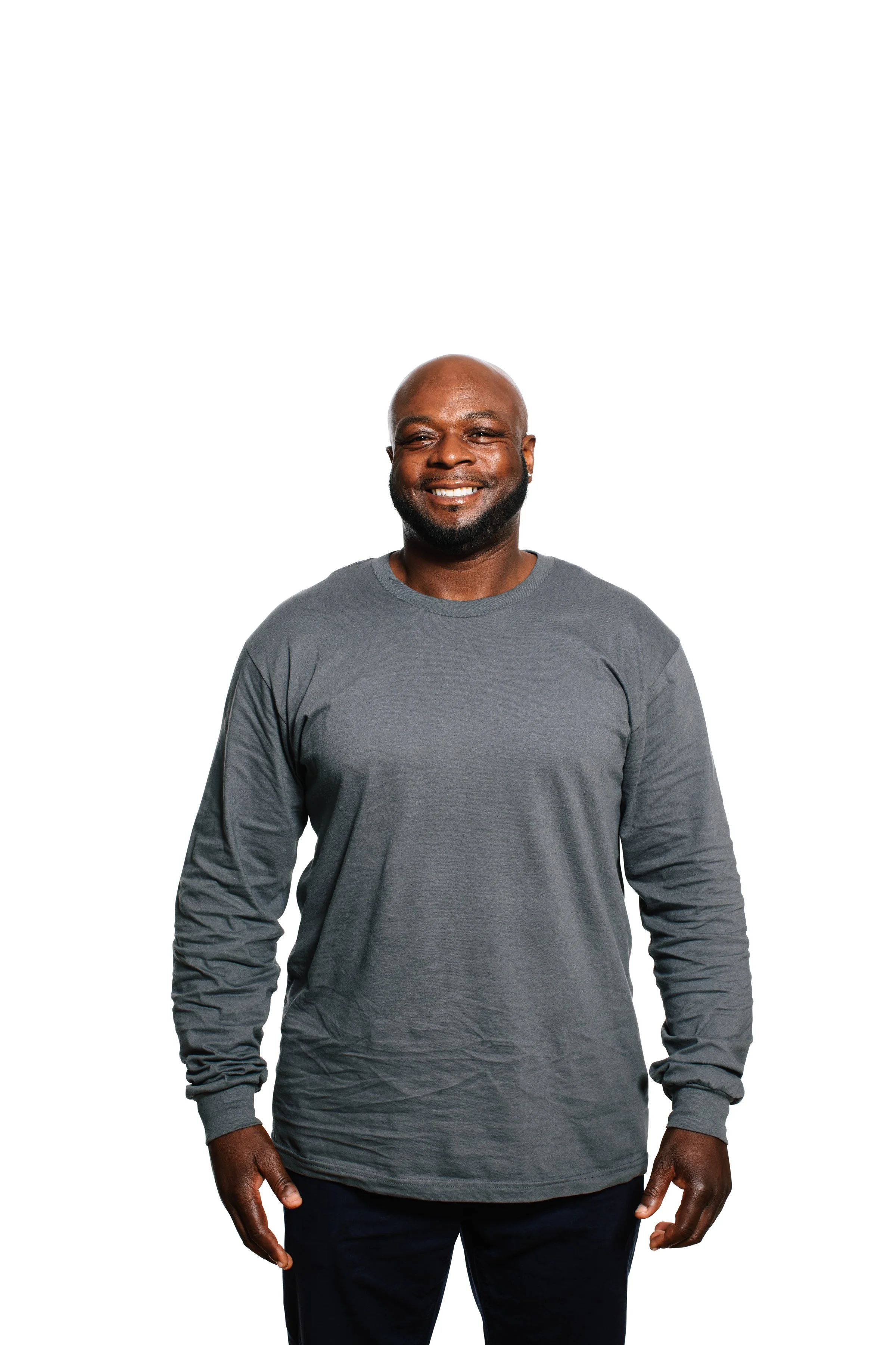 Smiling African American man with beard wearing a gray long-sleeve shirt and dark pants, standing against a white background.