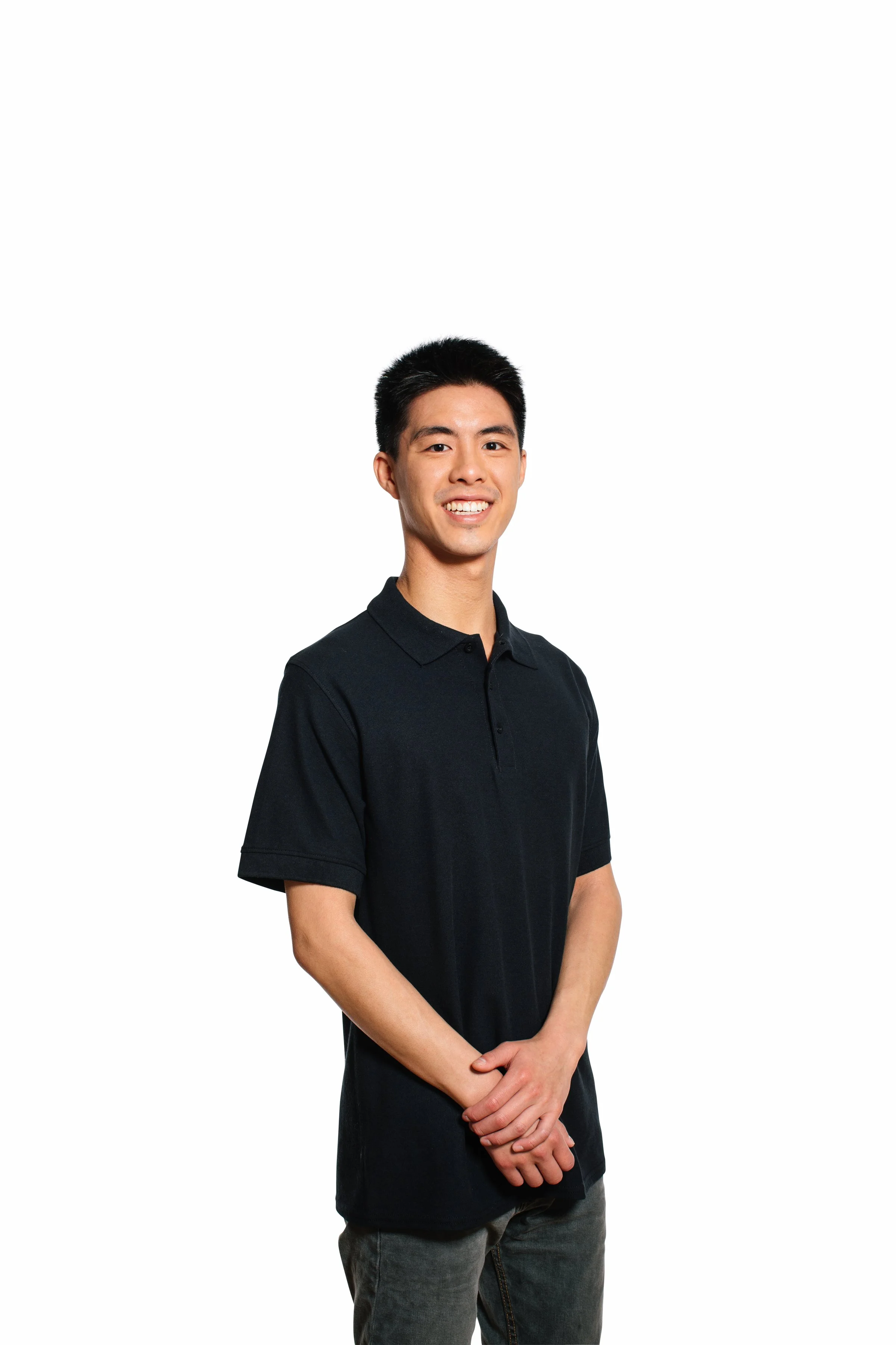 A young Asian man with short black hair, wearing a black polo shirt and gray jeans, standing with hands clasped in front, smiling against a plain white background.