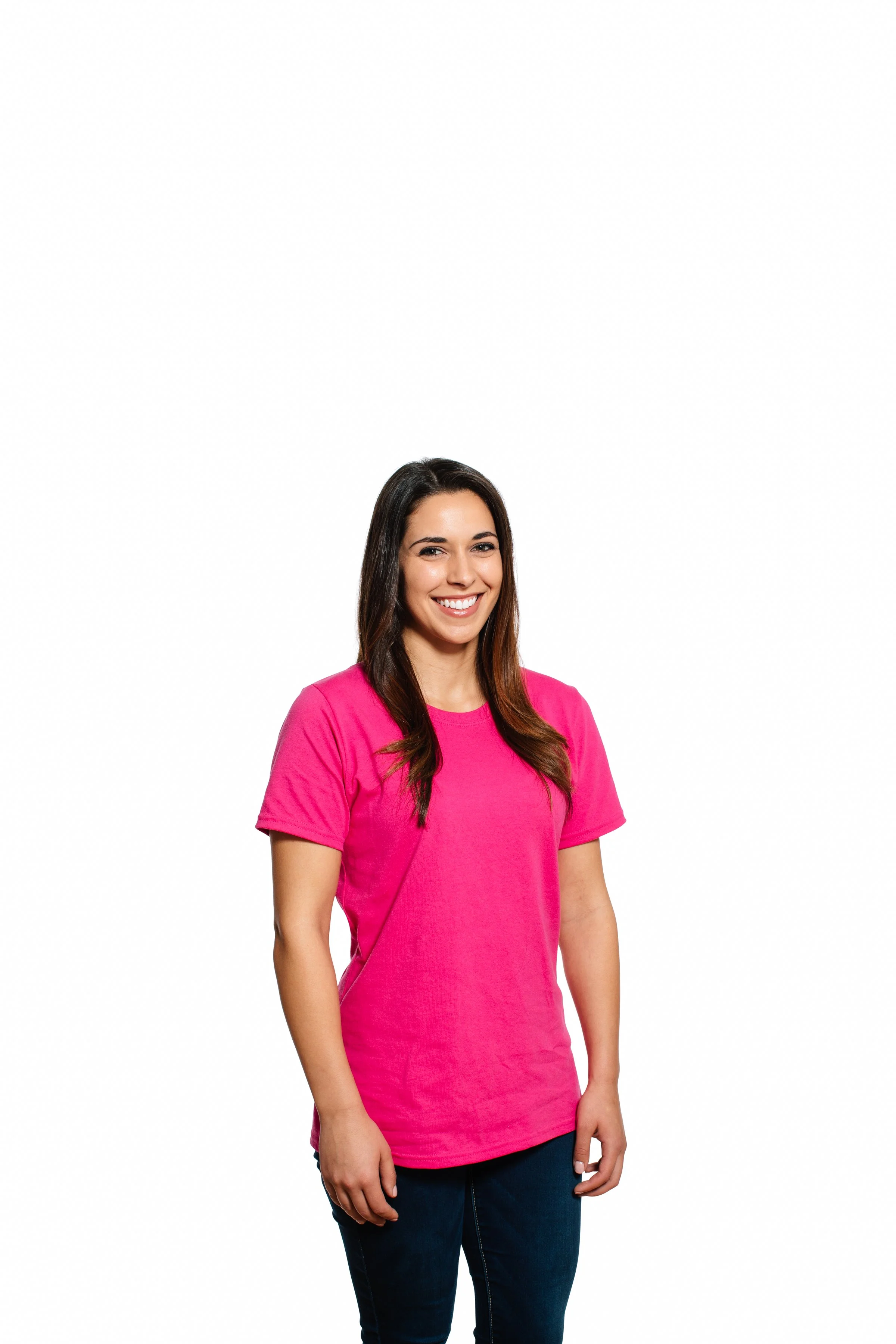 A woman with long dark hair smiling, wearing a bright pink t-shirt and dark jeans, standing against a plain white background.