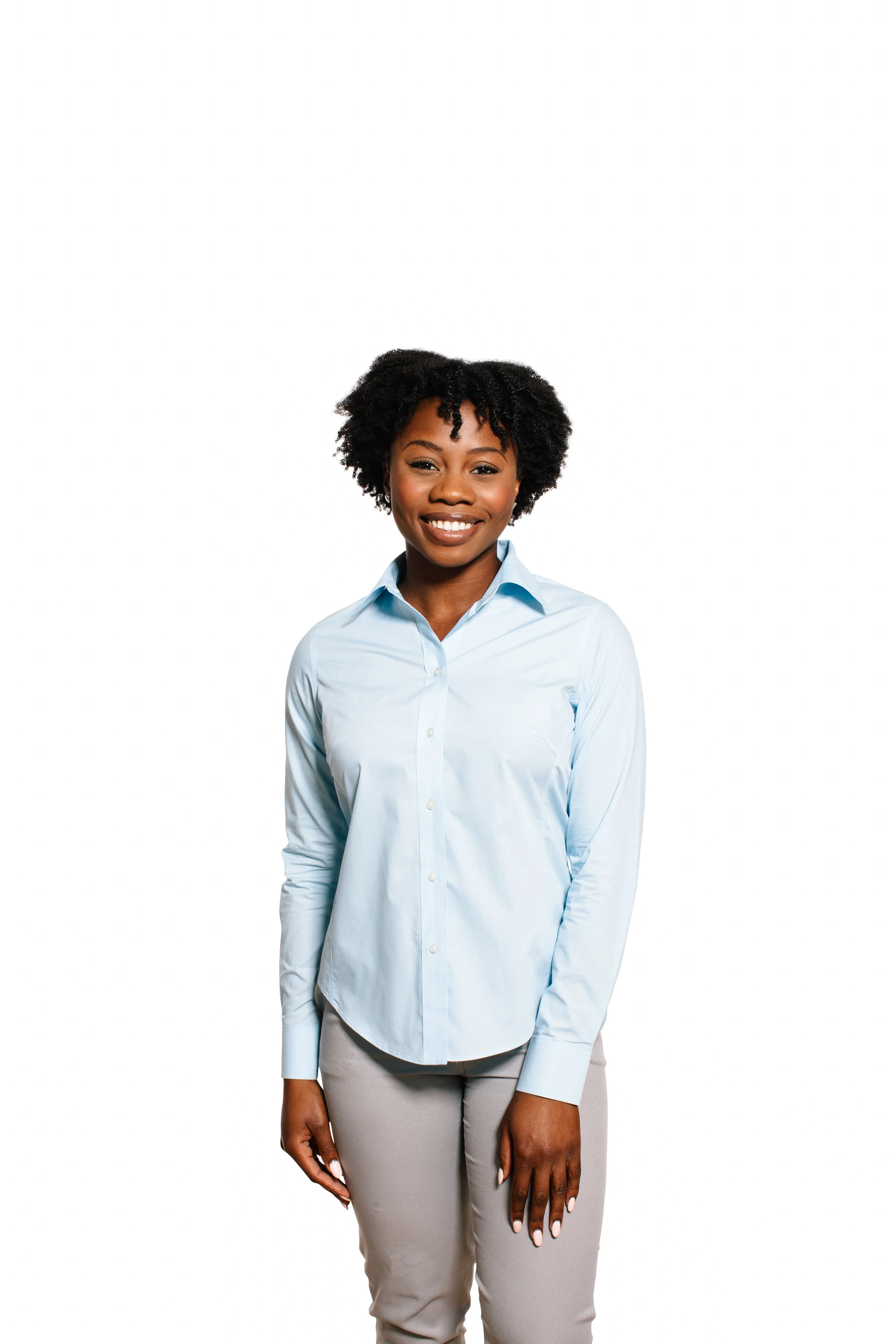 A woman with dark skin and natural curly hair, wearing a light blue button-up shirt and beige pants, smiling while standing against a plain white background.