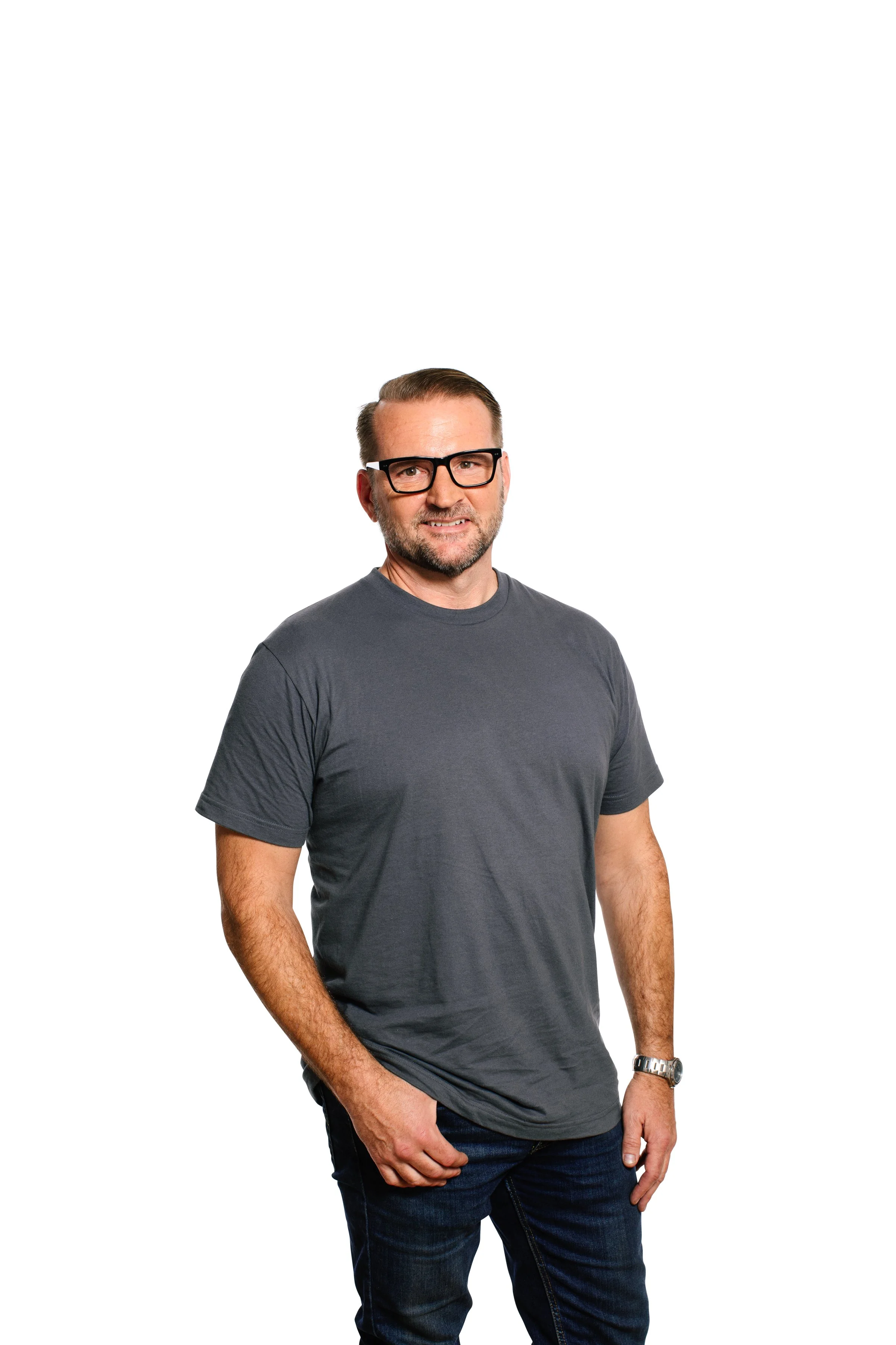 A middle-aged man with light skin, short brown hair, a beard, and glasses wearing a gray t-shirt and dark jeans standing against a white background.