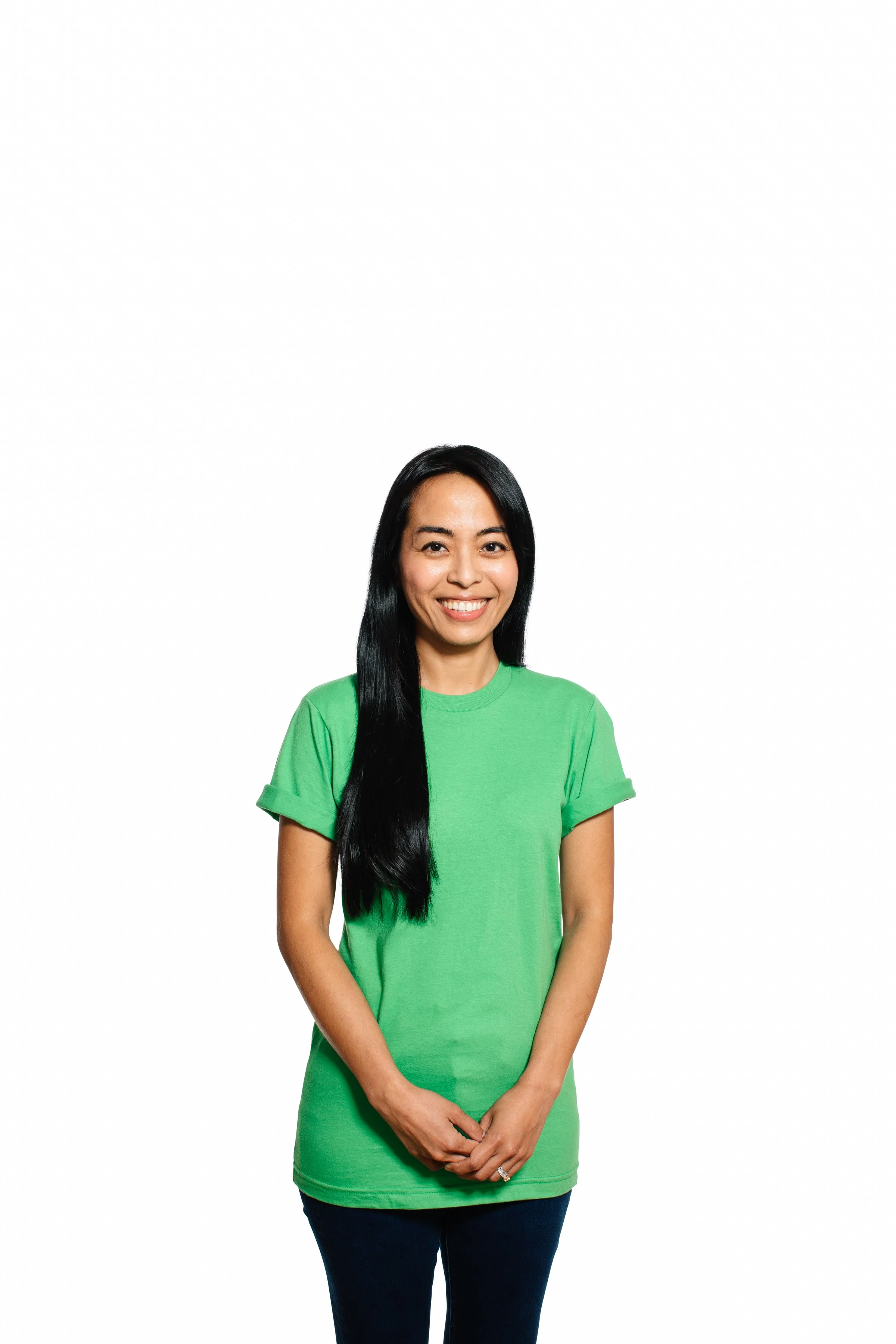 A woman with long black hair smiling, wearing a green T-shirt, standing with her hands clasped in front of her against a white background.