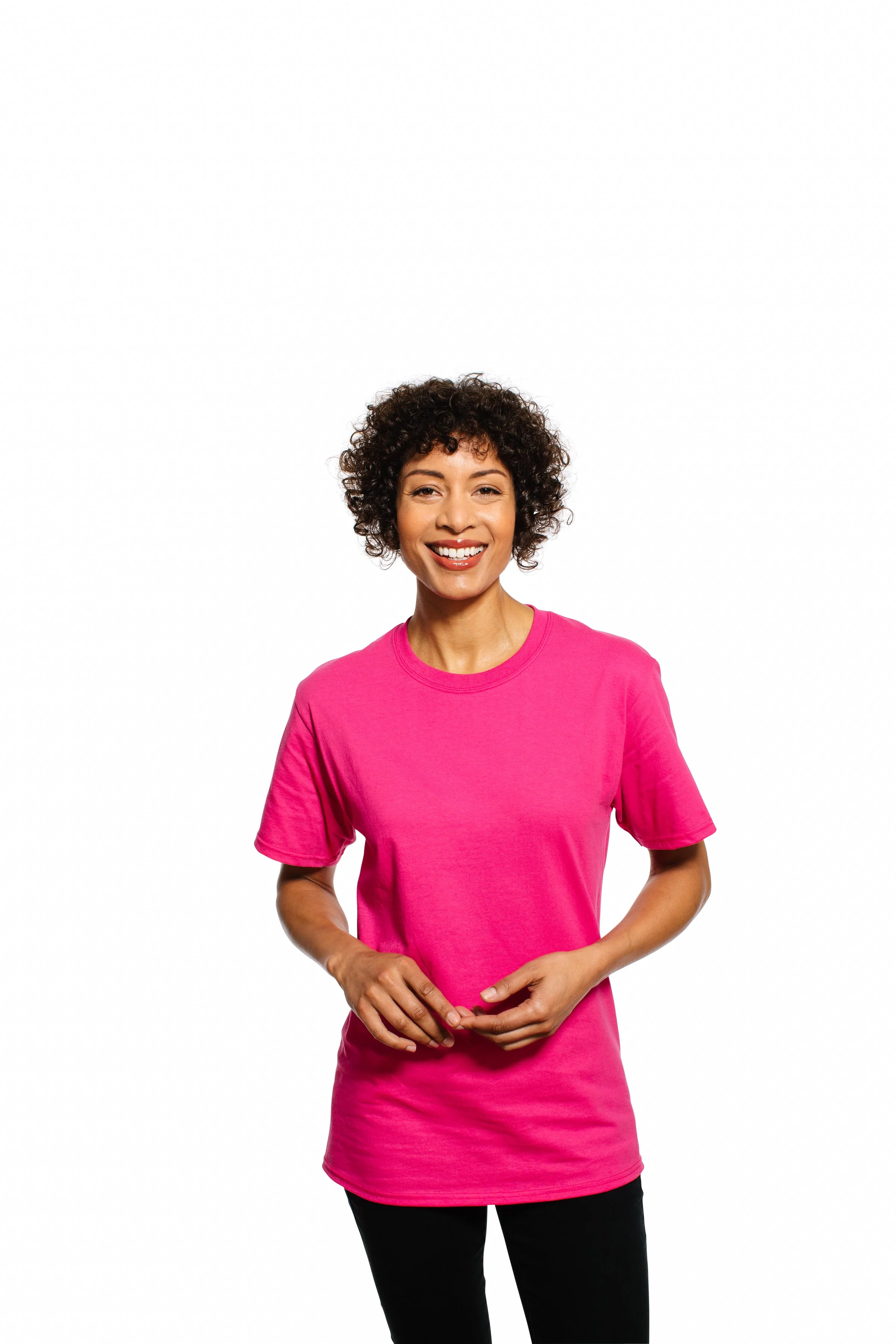 A smiling woman with curly hair wearing a pink T-shirt and black pants standing against a white background.