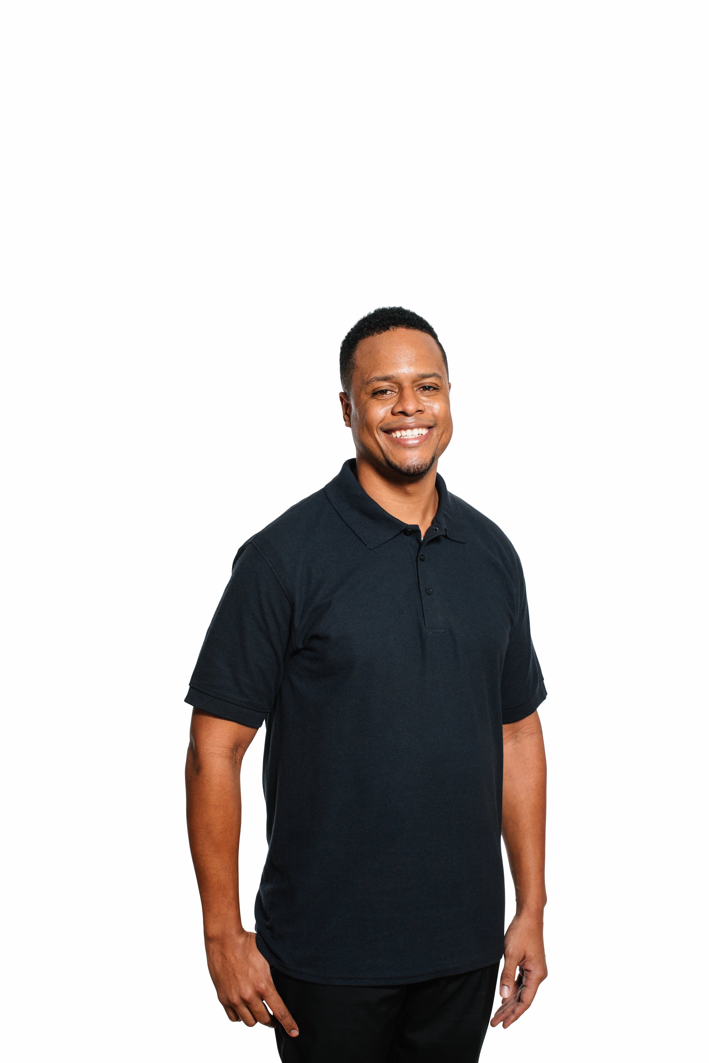A man with short curly hair and a goatee, smiling, wearing a dark polo shirt and black pants, standing against a white background.