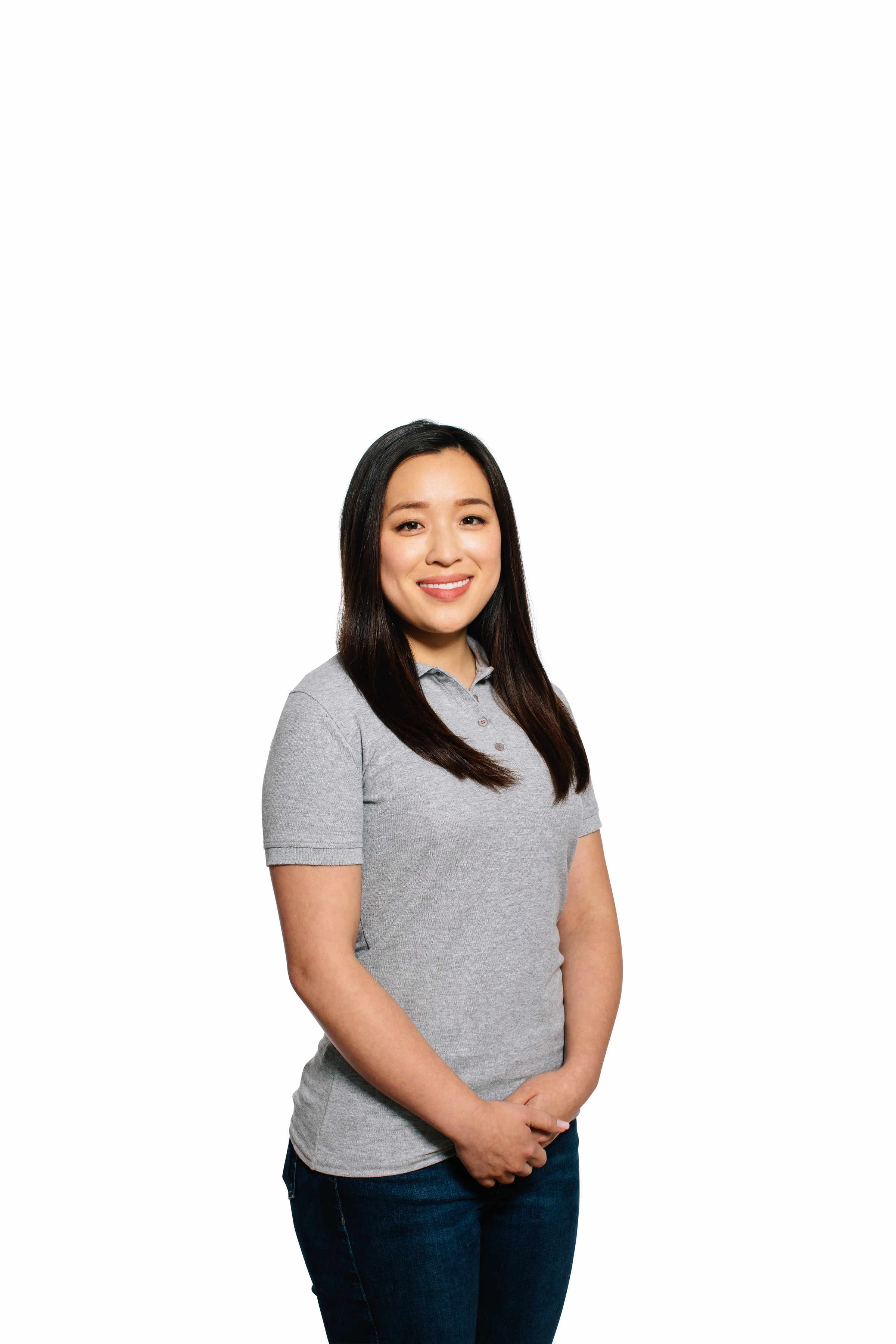 A woman with long dark hair, wearing a gray polo shirt and dark jeans, smiling and standing against a plain white background.