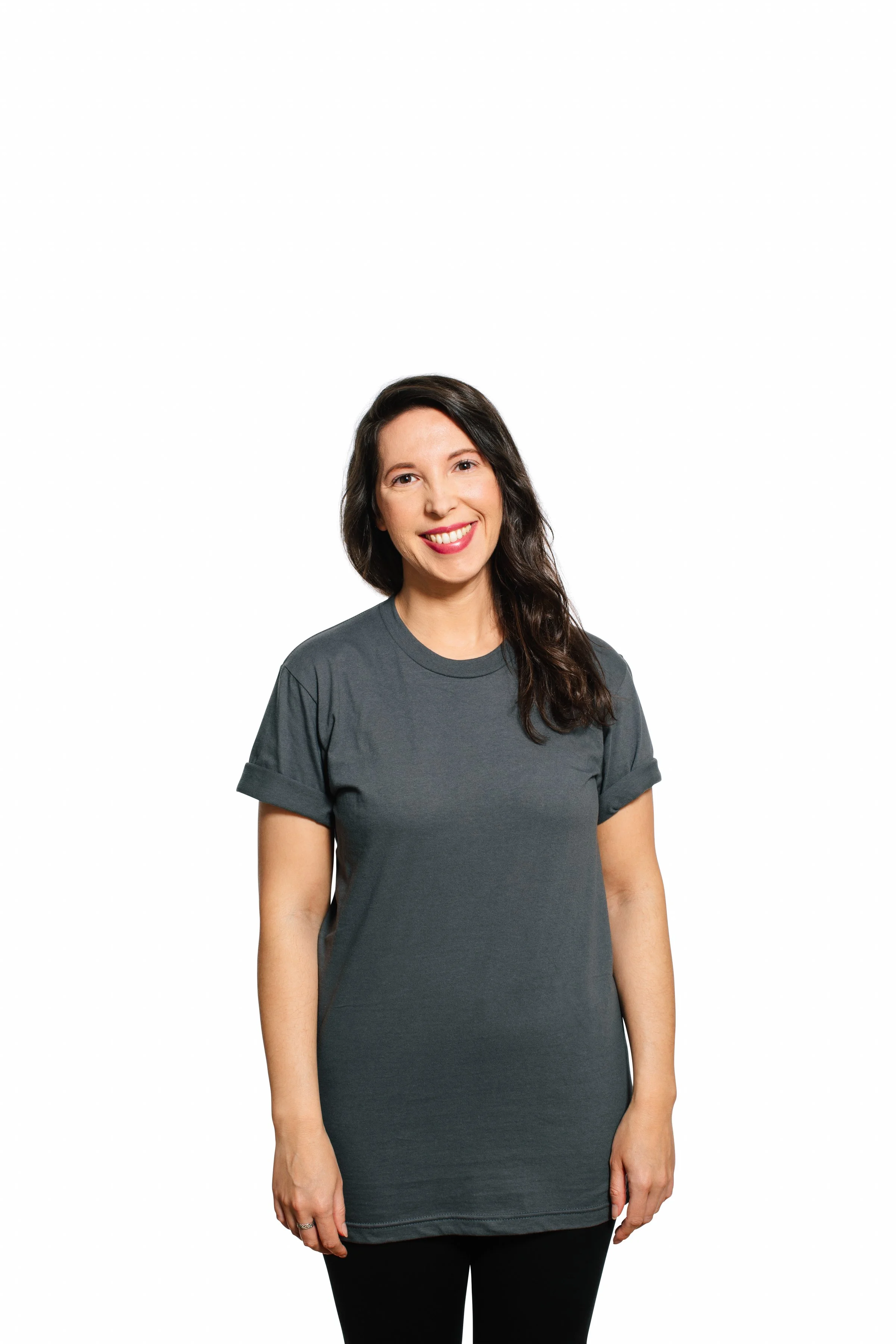 A woman with long dark hair smiling, wearing a gray T-shirt and black pants, standing against a white background.
